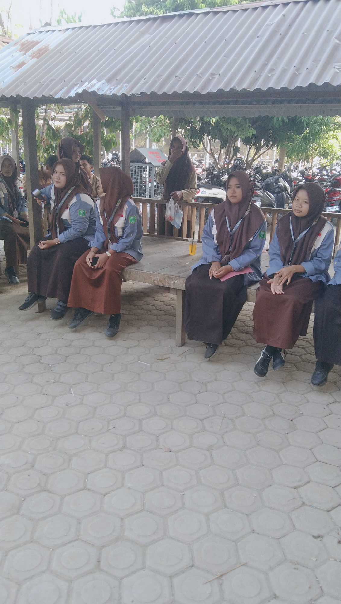 In Rhieng Blang, Indonesia, a group of young people, likely students, are gathered under a corrugated metal roof structure. Five individuals are clearly visible, seated on a wooden bench. They are wearing matching uniforms consisting of light blue shirts with emblems, brown headscarves, and long brown skirts. Their posture and expressions suggest they are waiting or observing. In the background, a variety of motorcycles are parked, indicating a public or school setting. The ground is paved with hexagonal concrete blocks. The overall atmosphere appears to be casual and outdoor.
