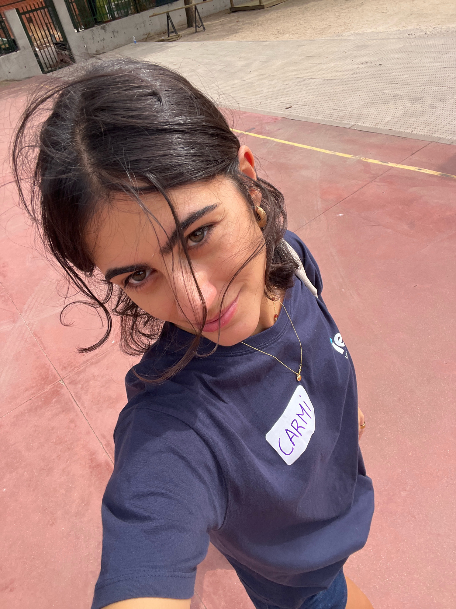 A young woman with dark hair, wearing a navy blue t-shirt with the name "CARMI" written in purple on a white nametag, is taking a selfie outdoors. Her hair is windswept and frames her face. She is wearing a delicate gold necklace and a small gold hoop earring. The background suggests an outdoor recreational area with a red-toned paved surface and a lighter-colored section of ground, possibly a sports court or park, with a yellow line marking a boundary. Behind her, a metal fence and some construction materials are visible, hinting at a location possibly under renovation or construction within the city of Getafe, Spain. The lighting indicates it is daytime.