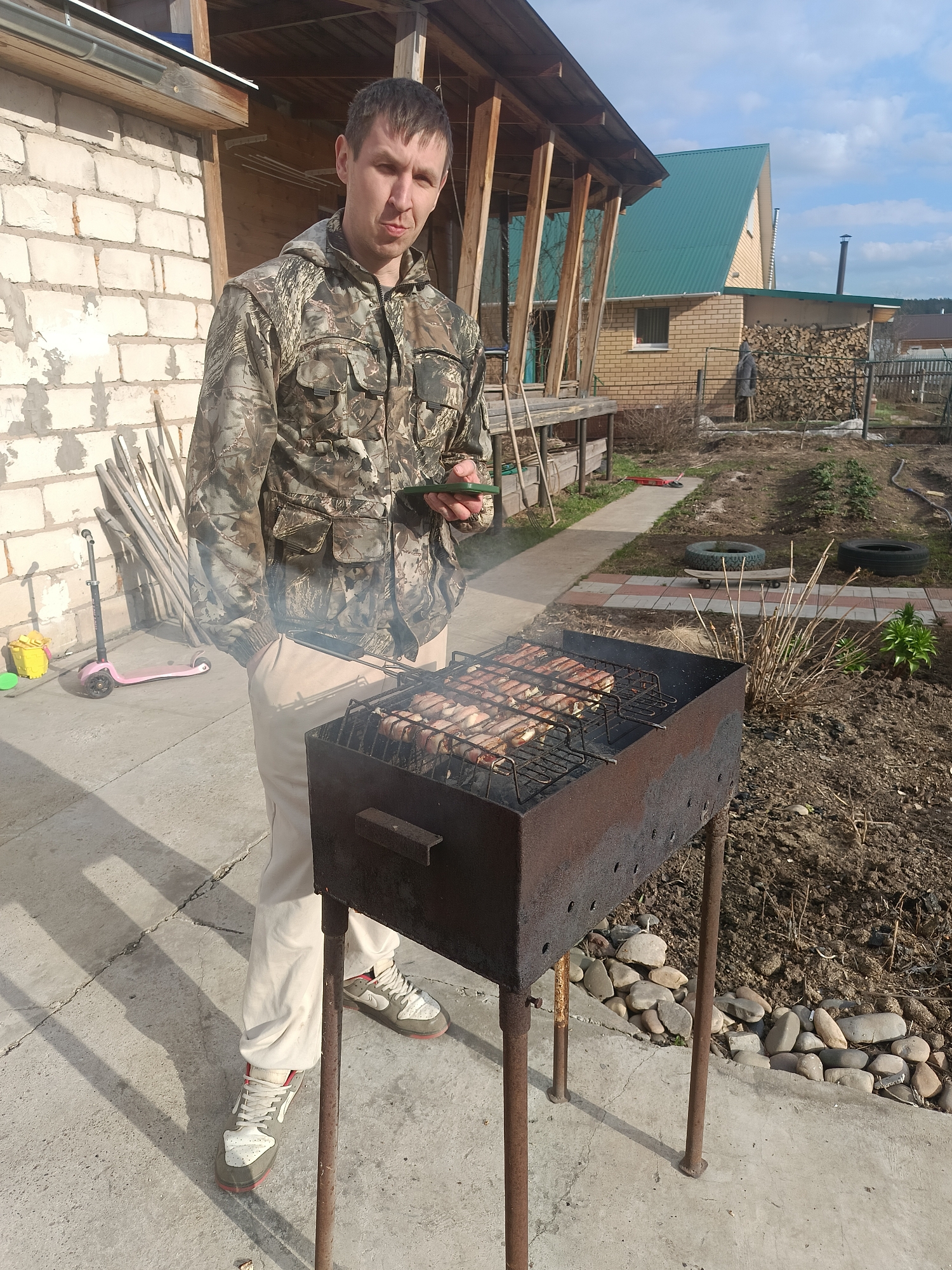 A man in a camouflage hooded jacket, light-colored pants, and grey and white sneakers stands by a rustic, dark metal grill (mangal) where pieces of meat are cooking on a grate, emitting smoke. He holds a green-cased smartphone in his left hand and looks at the camera with a neutral expression.
The setting is an outdoor area, likely a backyard or dacha in Chaikovsky, Russia. To the man's left, a weathered white brick wall with wooden supports is visible, against which lean a pink child's scooter, a yellow bucket, and several long wooden poles. The ground beneath him is a concrete slab.
Beyond the concrete, a paved path with red and grey tiles leads into a dirt area with dry, sparse vegetation, old car tires, and a wooden skateboard. Further in the background, a brick house with a steep, green corrugated metal roof and a chimney stands. Next to the house, a large stack of firewood is neatly piled. The sky is clear blue with a few scattered clouds, and the scene is brightly lit by direct sunlight, casting distinct shadows, indicating it is daytime, likely mid-morning or afternoon. No visible text is discernible in the image.