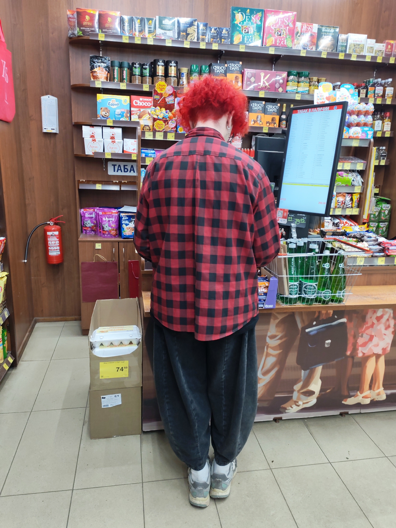 This image captures a person standing at the checkout counter of a small grocery store or convenience shop in Medvezhyegorsk, Russia. The individual, seen from behind, has striking bright red, curly hair and wears a red and black plaid shirt, dark baggy pants, and grey and white sneakers. They appear to be waiting to complete a purchase.
The setting is typical of a local Russian store, with wooden shelving stocked densely with various food and beverage items. To the person's left, a red fire extinguisher is mounted on the wall. Below it, cardboard boxes are on the tiled floor; one box contains several cartons of eggs and has a visible price tag of "74.69" rubles, along with the text "ЯЙЦО ПИЩЕВОЕ С1 ГОСТ 10 ШТ" (Table egg C1 GOST 10 pcs).
At the checkout counter, a metal shopping basket is filled with green-bottled beverages, possibly mineral water or soda, labeled "РЫЧАЛ-СУ" (Rychal-Su). A large vertical screen displays "ТАБАК В НАЛИЧИИ" (Tobacco in stock) and an "Итог" (Total) of "200.00" with other numbers like "997969", suggesting an ongoing transaction or inventory display.
Visible merchandise on the shelves includes various brands of tea (НУРИ ЦЕЙЛОНСКИЙ, JARDIN, RICHARD, MONARCH, DESERT TEA), snacks (Барни, ORION Choco, Chupa Chups, CHOCO DANS), chocolates (Адель, Raf), and pet food (Whiskas, Felix). A sign above one section clearly reads "ТАБА" (likely "ТАБАК" for tobacco). The front of the counter features a painted mural depicting the lower halves of people in casual attire. The store is lit by artificial lights, suggesting an interior space that could be operating at any time of day.