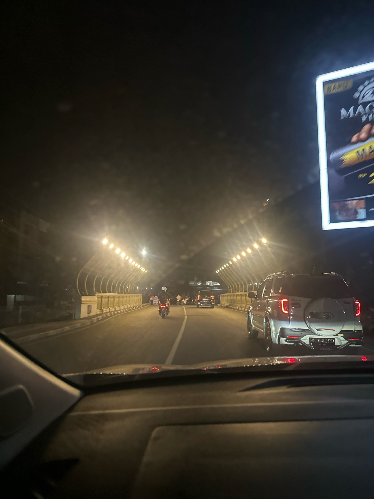 A night time street view from inside a vehicle in the city of Binjai, Indonesia. The road curves ahead, illuminated by a series of overhead lights. A white SUV is in the foreground, its brake lights illuminated, driving on the road. A motorcycle with a rider wearing a helmet is in the distance, also on the road. To the left of the road is a barrier with decorative golden posts, and beyond that, some buildings are visible. A large billboard with an advertisement is on the right side of the image, partially lit. The sky is dark.