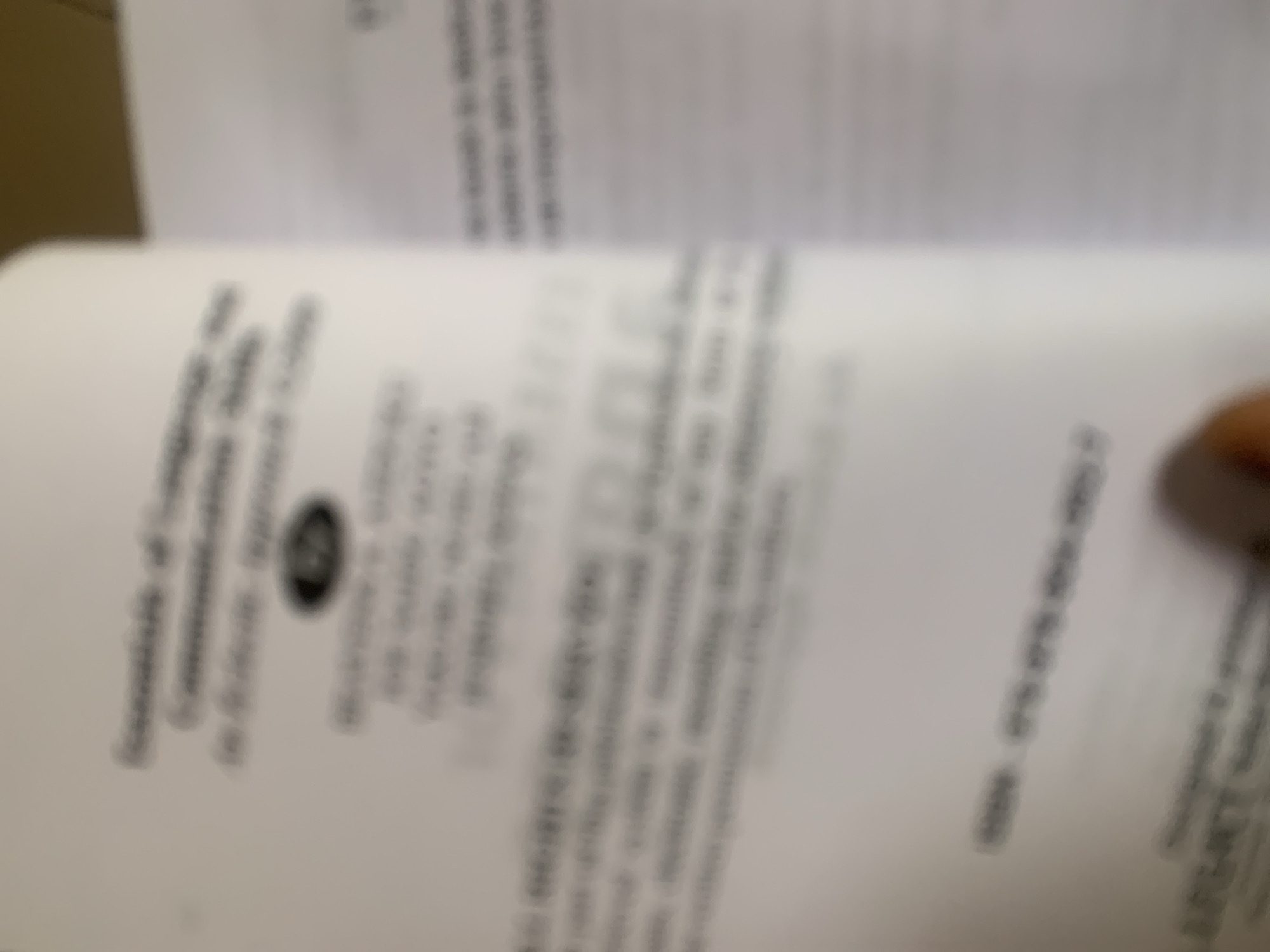 The image shows a blurred close-up of a book's title page. The text is largely illegible due to the blur, but some words appear to be "Essentials of Language and Communication Skills" and an emblem. Below this, there are more lines of text that are too blurry to decipher. Further down the page, a barcode-like graphic is visible. On the right side of the image, another page of the book is visible with blurred text. A finger is visible in the bottom right corner, obscuring a portion of the book. There is no information in the image that indicates a location in Jauro Yinu, Nigeria.