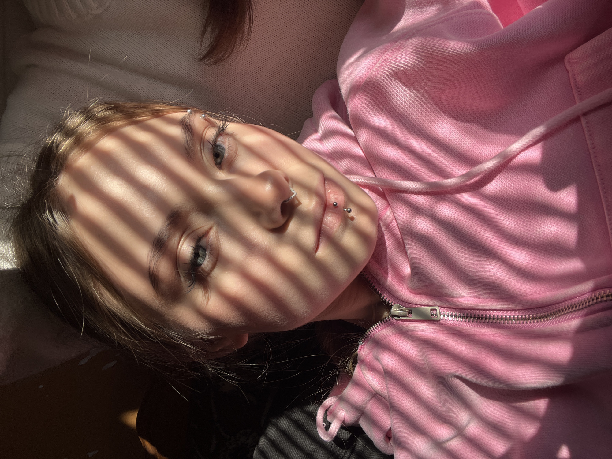 A young woman with light complexion, light hair, and light eyes is lying down. She is wearing a pink zip-up hoodie. The woman has multiple piercings, including one in her nose, two on her lower lip, and two on her eyebrow. Light from window blinds casts stark shadows across her face and the front of her hoodie. The environment and city of Фоки, Russia, cannot be confirmed.