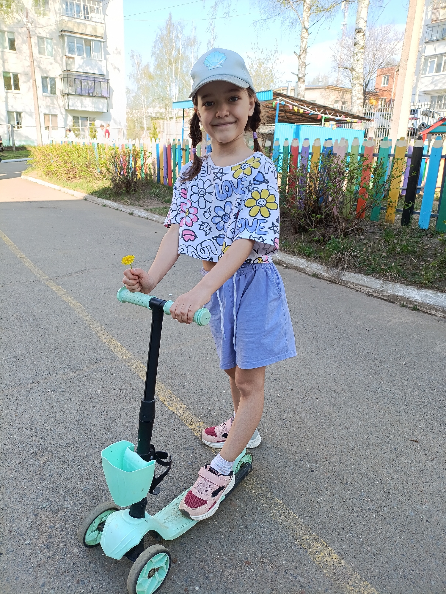A young girl stands on a light green three-wheeled scooter on a paved surface, likely a street or sidewalk, in Chaikovsky, Russia. She wears a light blue baseball cap, a cropped white t-shirt with a flower and "LOVE" print, and light purple shorts. She holds a small yellow flower in her left hand. Her hair is styled in pigtails.
Behind her is a colorful fence made to look like pencils. Beyond the fence, there are buildings and trees, typical of an urban environment. The scene is well-lit, suggesting it is daytime.