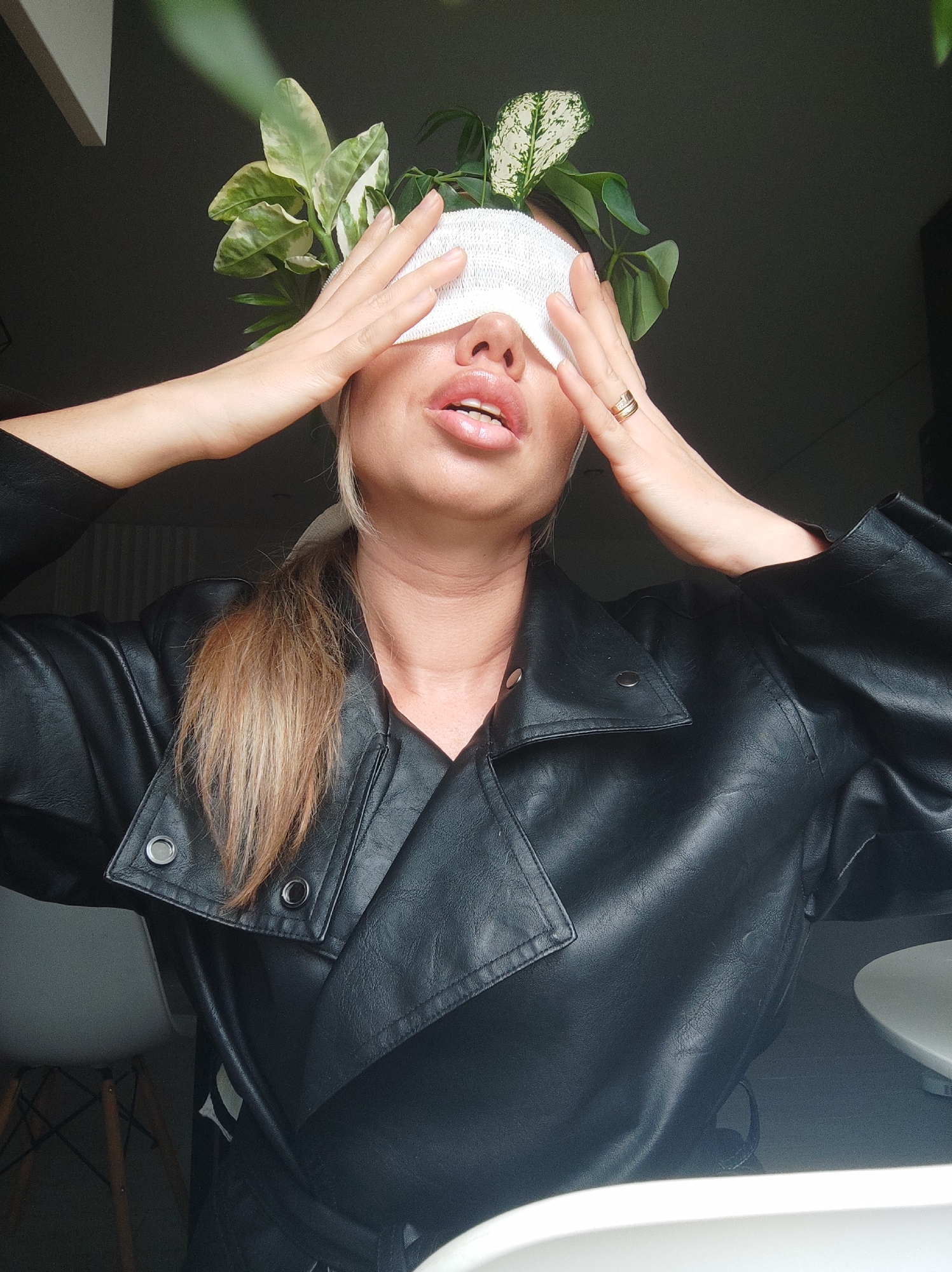 A woman is indoors, wearing a black leather jacket and a white bandage over her eyes, with green plants atop the bandage. She has blonde hair pulled back in a ponytail and is wearing a gold ring. Her hands are raised, touching the bandage. The location is unspecified; there is no information indicating it is in Votkinsk, Russia.