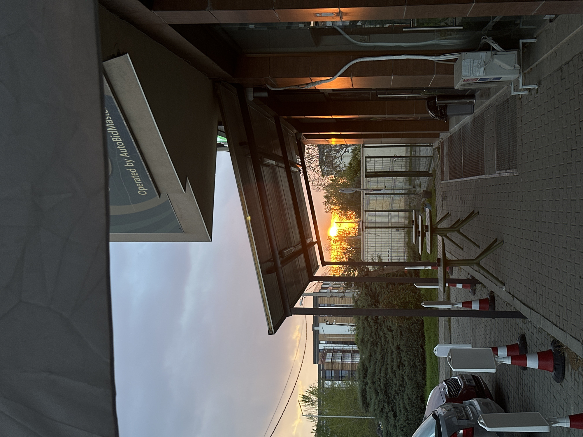 The image shows an outdoor setting, likely the drive-through area of a fast-food restaurant, possibly a McDonald's, in Plovdiv, Bulgaria. There are tables with seating under an awning, and a car is visible in the lower right corner. Traffic cones are placed along the edge of the road. The sky suggests either sunrise or sunset. The text "Operated by AutoBidMaster" is visible on the side of the building. An air conditioning unit is mounted on the wall. The sun is setting or rising in the distance through some trees.