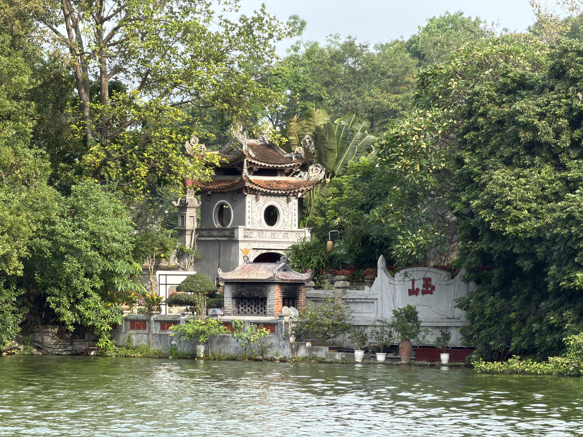 The image captures a view of the Temple of the Jade Mountain, located on Jade Island in Hoàn Kiếm Lake, Hanoi, Vietnam. The temple, a complex of structures with distinct architectural features, is partially obscured by lush greenery. The water of the lake reflects the sky and the surrounding vegetation. The temple itself features tiered roofs with ornate detailing and traditional designs. The building closest to the water has what appears to be a brick facade or chimney. A white wall with Asian characters written on it is to the right of the structure. Small potted plants decorate the perimeter of the island. The environment is serene and natural, with a backdrop of dense trees. There are no people visible in the scene.