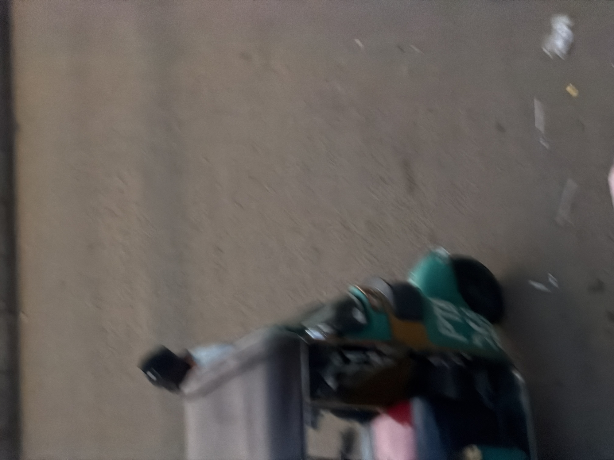 The image shows a blurry scene, possibly outdoors, with a predominantly neutral color palette. In the foreground, there is an object that appears to be a cart or a toy vehicle, with segments of green, yellow, and black visible. There is also a hint of pink and blue. Part of the object has some writing on it, but it is too blurred to decipher clearly. In the background, the surface is a dull, textured gray. Some small white specks are scattered across this surface. To the left of the scene, there is a vertical dark shadow. The overall impression is one of movement or a low-resolution photograph, making precise identification of objects and activities difficult. The image appears to be taken during daylight, but no specific time of day can be determined due to the blurriness and lack of distinct shadows or lighting cues. There are no clear indications of weather conditions. The setting appears to be an outdoor urban or semi-urban environment, potentially a street or an open area, given the textured ground and the presence of scattered debris. No identifiable people are clearly depicted, though there might be a blurred figure to the far right.
