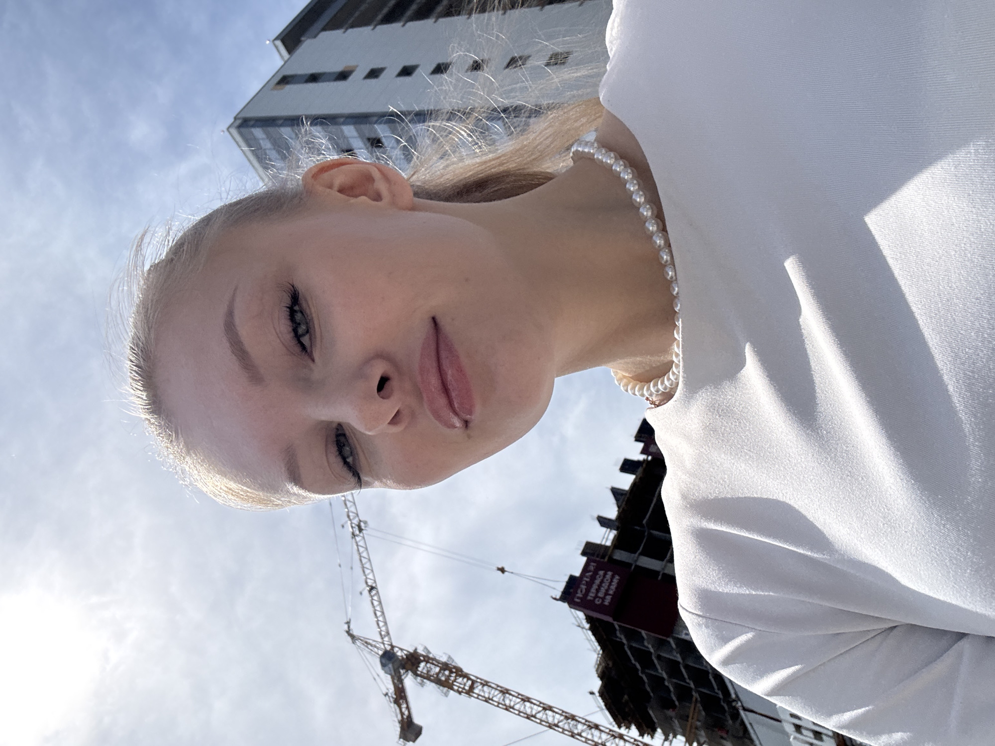 A close-up outdoor selfie depicts a young woman in an urban setting, likely Perm, Russia, captured on a bright, sunny day from a low angle looking upwards. The woman has fair skin, blonde hair, and light-colored eyes, with visible eyelashes and defined eyebrows. Her lips are slightly parted, revealing a small lip piercing on her lower lip. She is wearing a white top or blouse and a pearl necklace. Her expression is calm and neutral.
In the background, under a clear blue sky with scattered white clouds, several structures are visible. To the upper right, a modern, light-colored high-rise building with numerous windows stands. A large yellow construction tower crane dominates the middle-left background, indicating an active building site. Towards the lower right, a dark, partially constructed concrete building is visible. On this building, a red sign with white Cyrillic text reads "ПРОДАЖИ ТЕРРАСЫ С ВИДОМ НА КАМУ," which translates to "Sales of Terraces with a View of the Kama," referencing the Kama River in Perm. Bright, direct sunlight illuminates the scene, creating strong highlights on the woman's hair and clothing, suggesting it is midday or early afternoon.