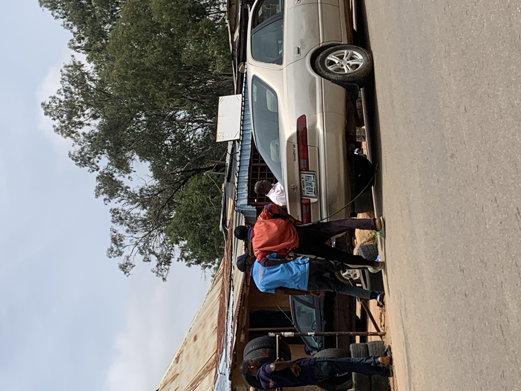 This image depicts an outdoor scene in Jos, Nigeria, likely during daylight hours under a cloudy sky. The primary subjects are several individuals gathered around a beige sedan. The scene appears to be a makeshift roadside service area.
In the foreground, several people are engaged in activity related to the car. One person, wearing a blue t-shirt, seems to be operating a device connected to the car, possibly inflating a tire. Another person, in an orange jacket, stands nearby. Further down, another individual in a dark t-shirt and jeans is visible, appearing to be resting or working on something near a stack of tires.
The setting includes a weathered, corrugated metal structure that serves as a shelter or shop. Tires are stacked around the area, indicating a tire repair or sales business. The car itself is a sedan, with its license plate visible, reading "YAB-347PN". The surrounding environment is somewhat rustic, with trees visible in the background and a paved road to the right of the image. The overall impression is one of a busy, practical, and possibly informal commercial operation.