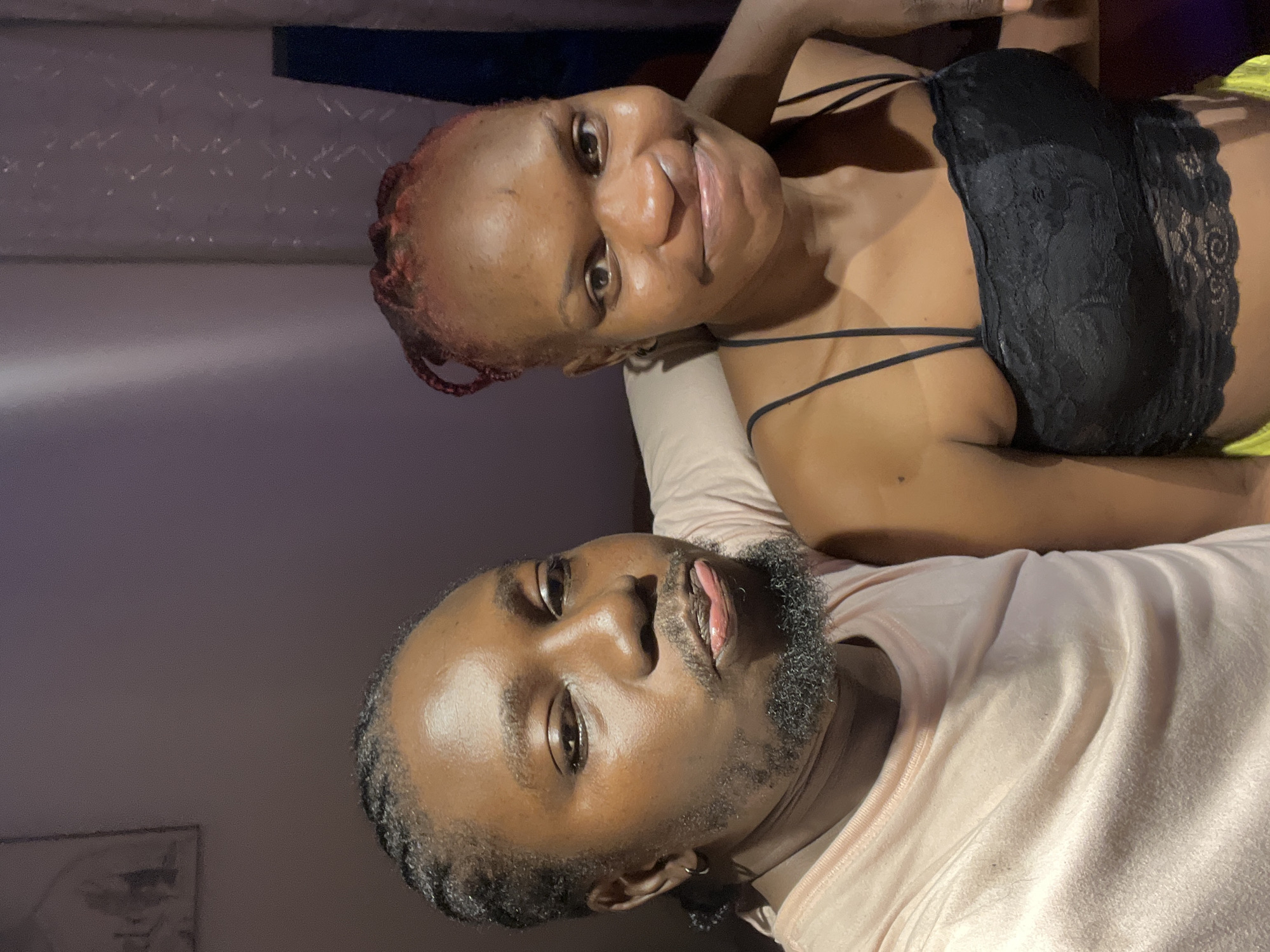 This is a selfie of two Black individuals, a man and a woman, posing closely together. The man, on the left, has braided hair, a goatee, and is wearing a light pink t-shirt. He is sticking his tongue out slightly. The woman, on the right, has reddish-brown braided hair and is wearing a black lace camisole. She is smiling slightly. They appear to be indoors, with a wall and curtains visible in the background. The lighting suggests it is evening or nighttime. There is no specific indication of the location being in Jos, Nigeria, within the image itself.