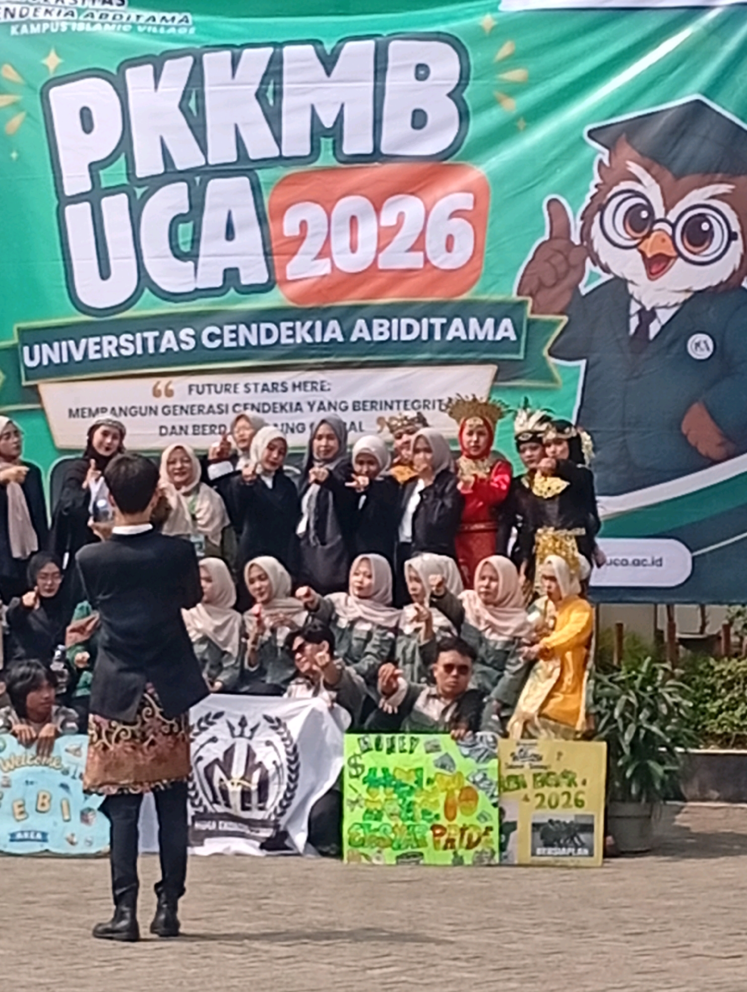 This image captures an outdoor event, likely an orientation or welcome ceremony, at Universitas Cendekia Abbidtama in Kelapa Dua, Indonesia. The central focus is a large green banner with bold white lettering that reads "PKKMB UCA 2026," indicating the event's purpose and year. To the right of the banner, a cartoon owl wearing a graduation cap and gown gestures with its thumb up, serving as a mascot.
Below the banner, a group of students, predominantly young women wearing headscarves and various attire, are posing for a photograph. Some are dressed in traditional outfits, including a woman in a red and gold outfit and another in a black outfit with gold embellishments. A young man in a black suit and traditional patterned skirt stands in the foreground, facing away from the camera, possibly addressing the group or participating in the ceremony.
To the left and right of the students, several colorful signs are visible. One sign on the left reads "Welcome" and "EB1 AREA" in blue and yellow. Another on the right is yellow and features text that includes "SELAMAT DATANG," "UCA BG R. 2026," and a photo of four people. Behind the students and signs, there are potted plants and what appears to be fencing or a wall. The ground surface is paved, suggesting an outdoor plaza or courtyard. The overall atmosphere appears festive and celebratory.