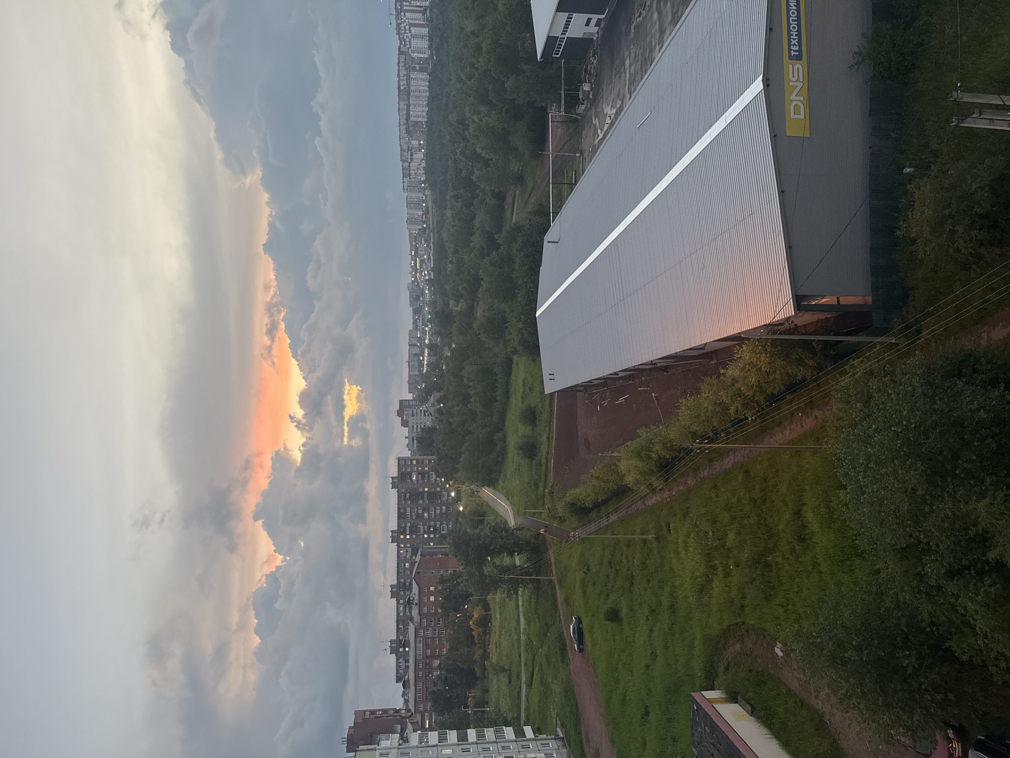 The image depicts an aerial view of a suburban or industrial area in Bratsk, Russia, at dusk. The sky is filled with dramatic, textured clouds, with a warm orange glow of sunset visible through a gap in the clouds, creating a striking contrast with the cooler, gray tones of the rest of the sky.
In the foreground, a large, modern warehouse-style building with a corrugated metal roof dominates the right side of the frame. A yellow sign with blue lettering, reading "DNS ТЕХНОПОИНТ", is affixed to the side of the building. A low green fence runs along the perimeter of the building.
To the left of the warehouse, a wide expanse of green grass stretches towards a dirt path. A dark-colored sedan is parked on this path. Further in the distance, a cluster of trees forms a dense green belt across the middle ground.
Beyond the trees, a row of multi-story apartment buildings stretches across the horizon. Many of these buildings have illuminated windows, indicating that it is evening. The architecture suggests a typical Soviet-era housing development. In the far distance, more buildings and structures are visible, hinting at the larger urban landscape of Bratsk.
The overall impression is one of an urban environment at the edge of nature, with the imposing structures of industry and housing set against a backdrop of a vibrant, atmospheric sky. The presence of the DNS sign suggests a commercial or retail area within this industrial or suburban zone.