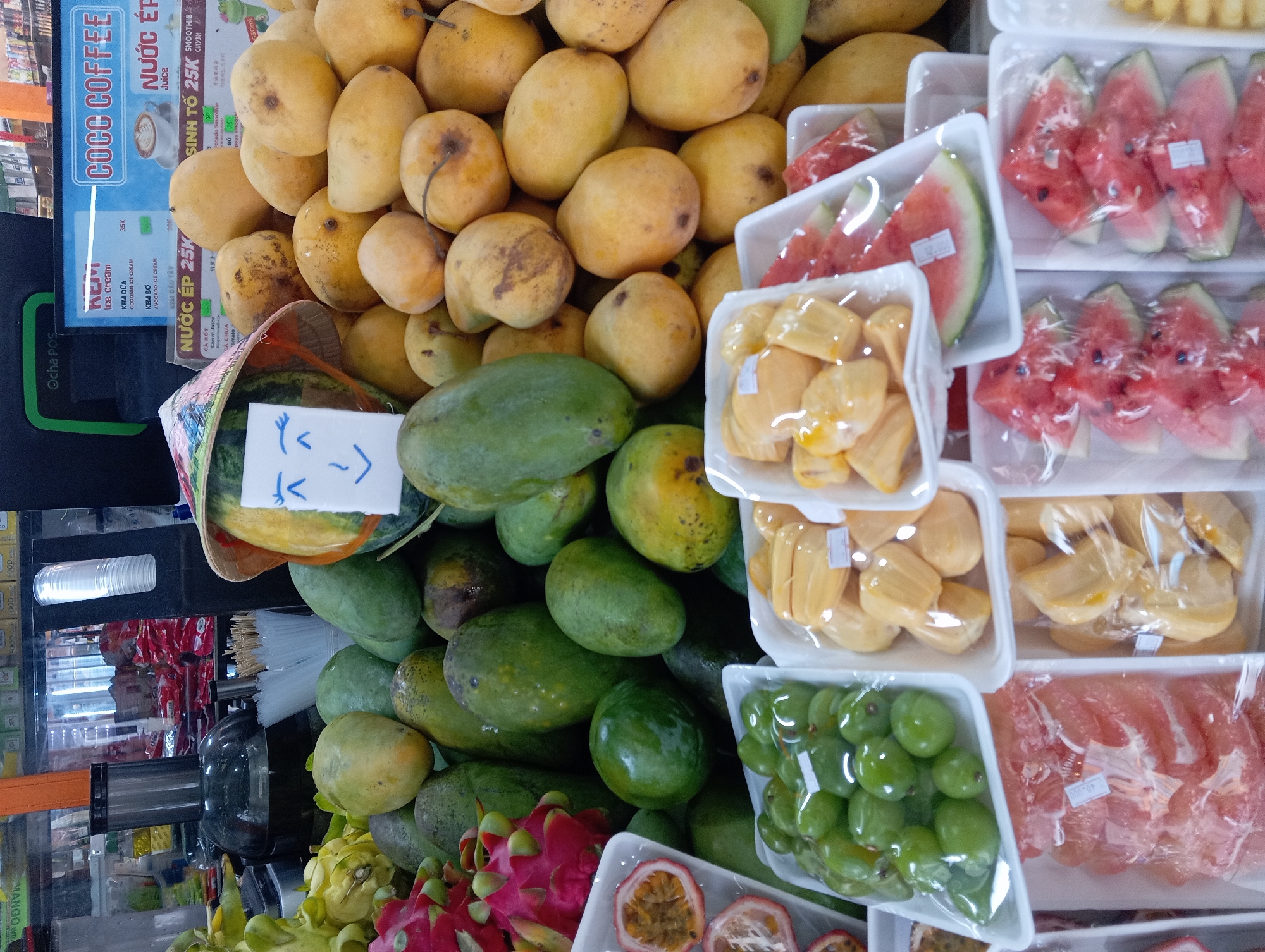 In Nha Trang, Vietnam, a vibrant market stall showcases an abundant array of fresh fruits. Piles of yellow and green mangoes dominate the display, interspersed with dragon fruit, passion fruit halves, green grapes, and slices of jackfruit and watermelon, all neatly arranged in plastic containers. A decorative watermelon, adorned with a hand-drawn smiley face on a paper sign and a traditional conical hat, stands out as a focal point. In the background, a juice dispenser and a variety of packaged goods suggest a bustling commercial environment. The scene captures a typical street-side fruit vendor, offering a colorful and enticing selection to passersby.