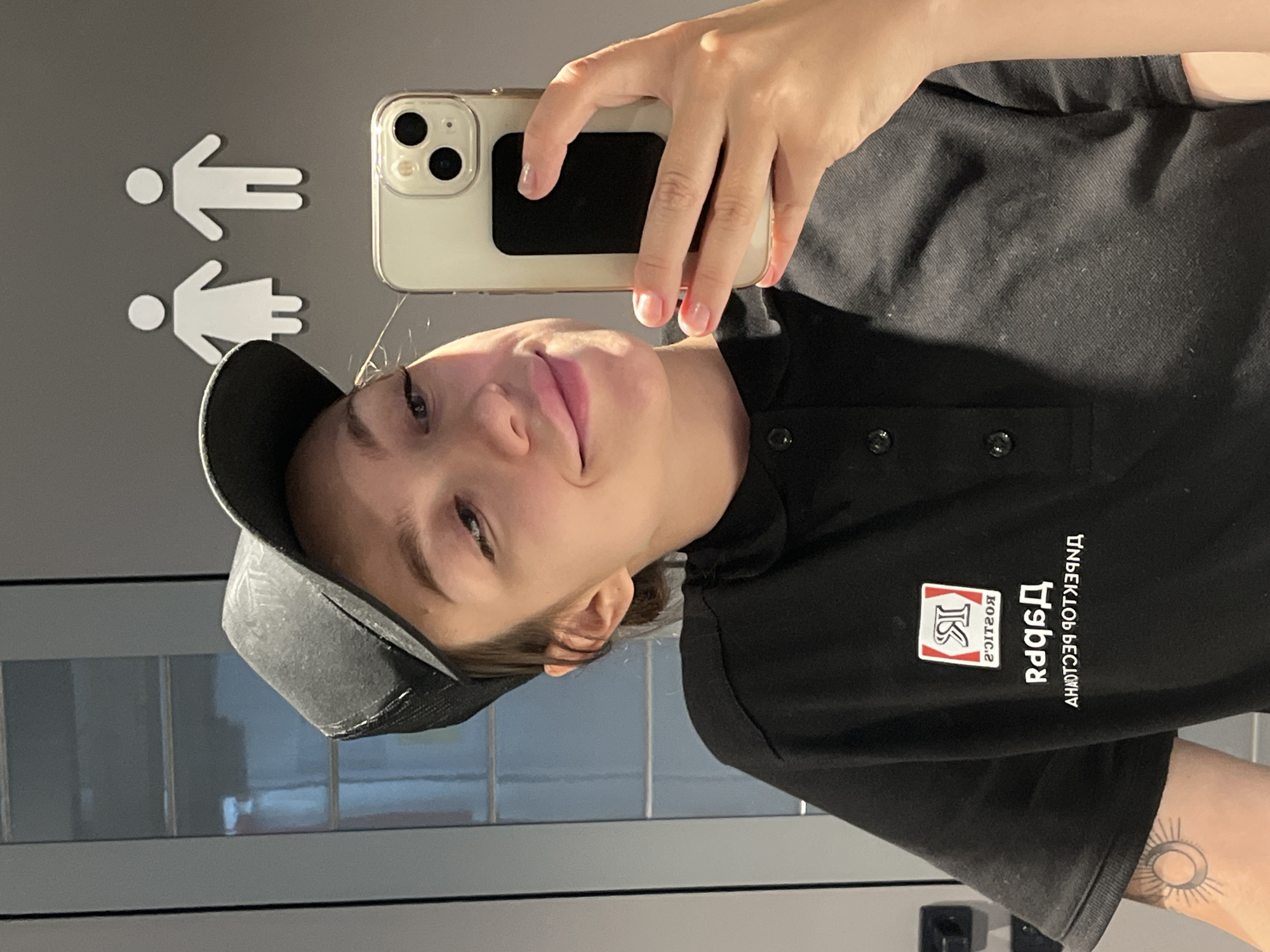 A young woman wearing a black baseball cap and a black polo shirt is taking a selfie in a restroom. The polo shirt has a logo on the left chest area with Cyrillic lettering. Her left arm, visible from the shoulder down, has a tattoo of a sun and crescent moon. Behind her, on the wall, are white male and female restroom symbols. The environment appears to be a public restroom, possibly in Chaikovsky, Russia, given the Cyrillic lettering on her shirt and the general aesthetic. She is holding a smartphone in her right hand to capture the photo.