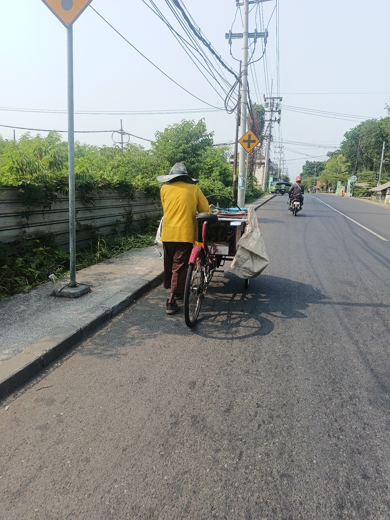 In Sruni, Indonesia, a person wearing a wide-brimmed hat and a yellow shirt walks along a paved road, pushing a red and silver tricycle. The tricycle has a cargo area loaded with two large sacks, one appearing to be made of woven material and the other a lighter colored sack. The person is dressed in brown pants and dark shoes. They are walking on the edge of the road, next to a sidewalk and a gray metal fence. Behind the fence, dense green foliage is visible. Overhead, a complex network of power lines crisscrosses the sky, supported by utility poles. A yellow road sign with a black cross symbol is mounted on one of the poles. Further down the road, other vehicles are present, including a green car and a motorcycle with a rider. The road is asphalt and appears to be in good condition. The scene is illuminated by natural daylight.