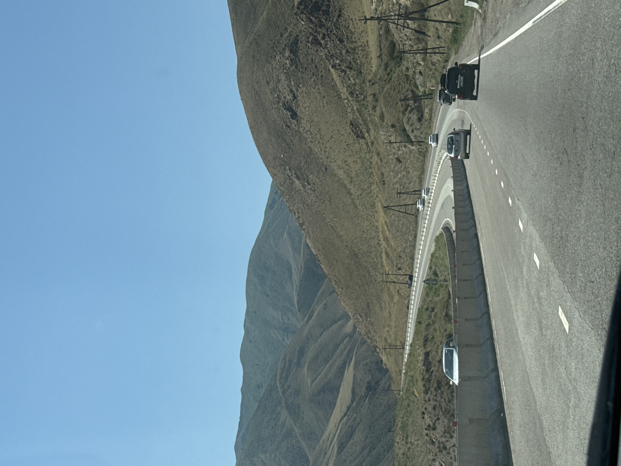 The image depicts a scenic view of a highway winding through mountainous terrain under a clear blue sky. The road, asphalted and with white lane markings, curves to the left, bordered by a concrete barrier on the outer edge. Several vehicles are visible on the road, including a white SUV stopped behind the barrier, a silver sedan, and further ahead, a black SUV, a silver sedan, and two white sedans. The surrounding landscape is characterized by dry, grassy hills with undulating contours, some with rocky outcrops. Utility poles with wires are strategically placed along the roadside. The photograph appears to be taken from a moving vehicle, looking forward. The location is identified as the vicinity of Cholok, Kyrgyzstan.