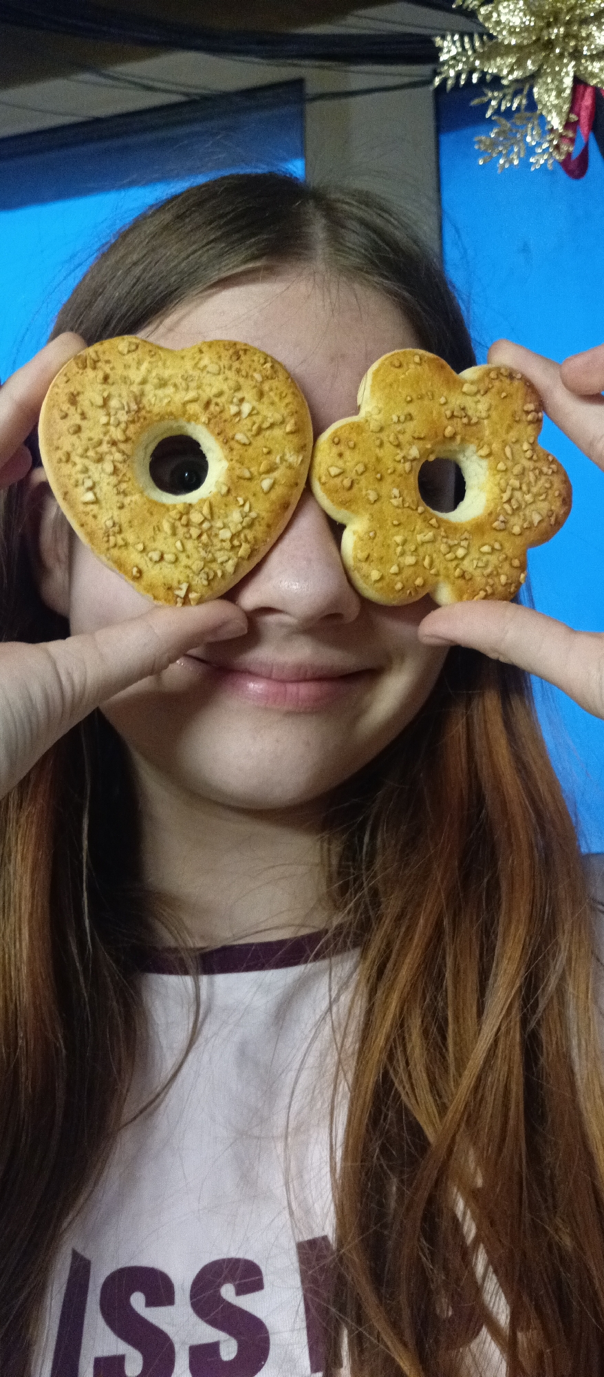 A medium close-up shot features a young person, likely female, with fair skin and long, reddish-brown hair, engaged in a playful activity. The person holds two light brown cookies, one heart-shaped and one flower-shaped, directly in front of their eyes, looking through the central holes in each. Both cookies are generously topped with small, chopped nut pieces, giving them a textured appearance. The person's fingers are visible, carefully positioning the cookies. A gentle, subtle smile is visible on their face, conveying a sense of amusement or happiness.
The background is a vibrant, solid blue, hinting at an indoor setting. In the top right corner, a glimpse of a shiny, golden, star-shaped or floral decoration, possibly a Christmas ornament, is visible, adding a festive touch. The person is wearing a white t-shirt, and part of a word or phrase, specifically the letters "ISS", can be seen on the fabric below their chin, partially obscured by their hair and the bottom edge of the frame. The image captures a lighthearted moment of fun.
