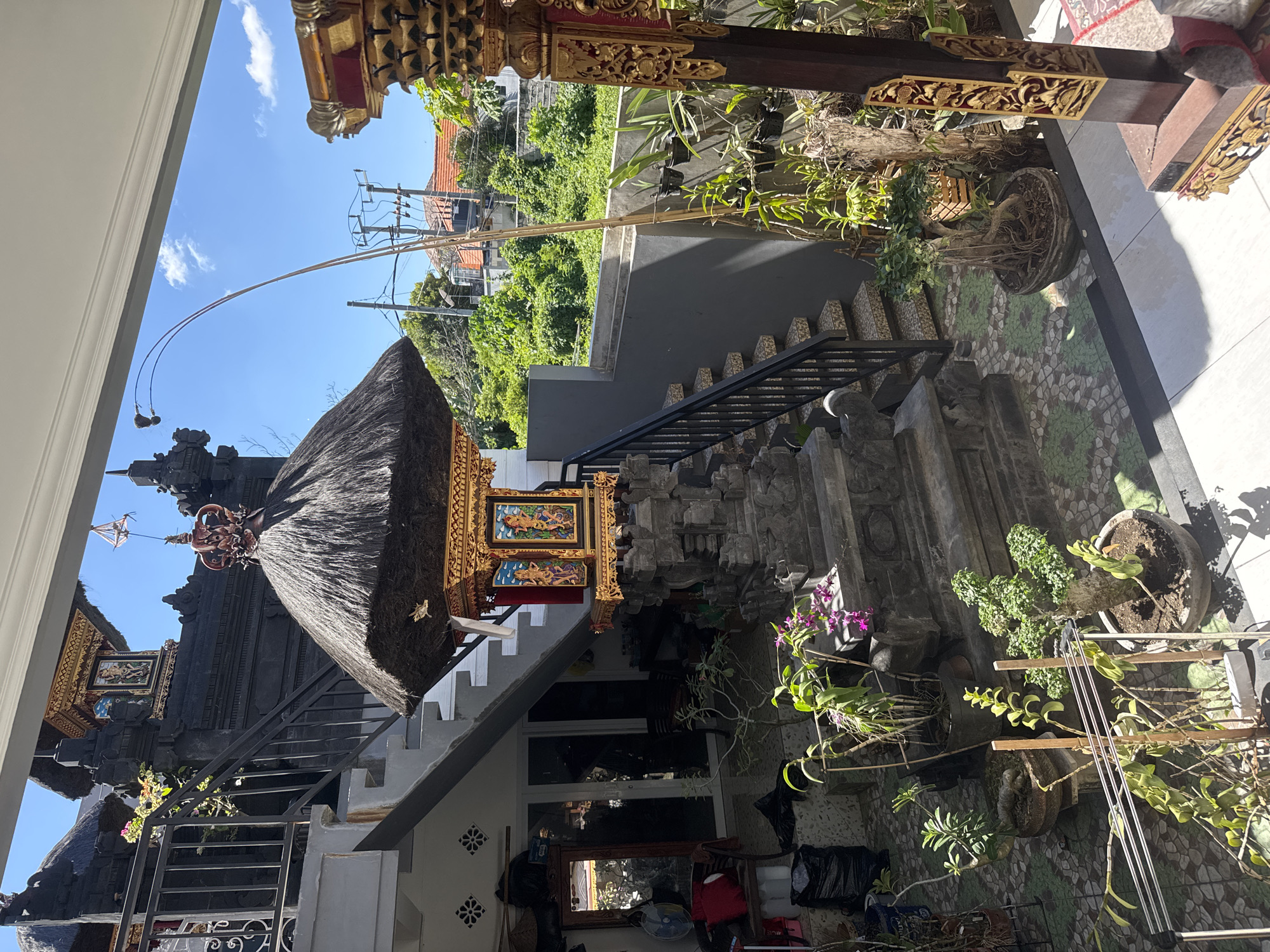The image shows a yard in Kuta, Indonesia, with a traditional Balinese temple or shrine as a focal point. The shrine is built with a thatched roof and ornate gold trim. The surrounding yard features stone steps, potted plants, and decorative stonework. A bamboo pole is visible extending into the sky. In the background, there's a view of rooftops and greenery, suggesting an urban or suburban residential area. The scene appears to be taken during a sunny day.