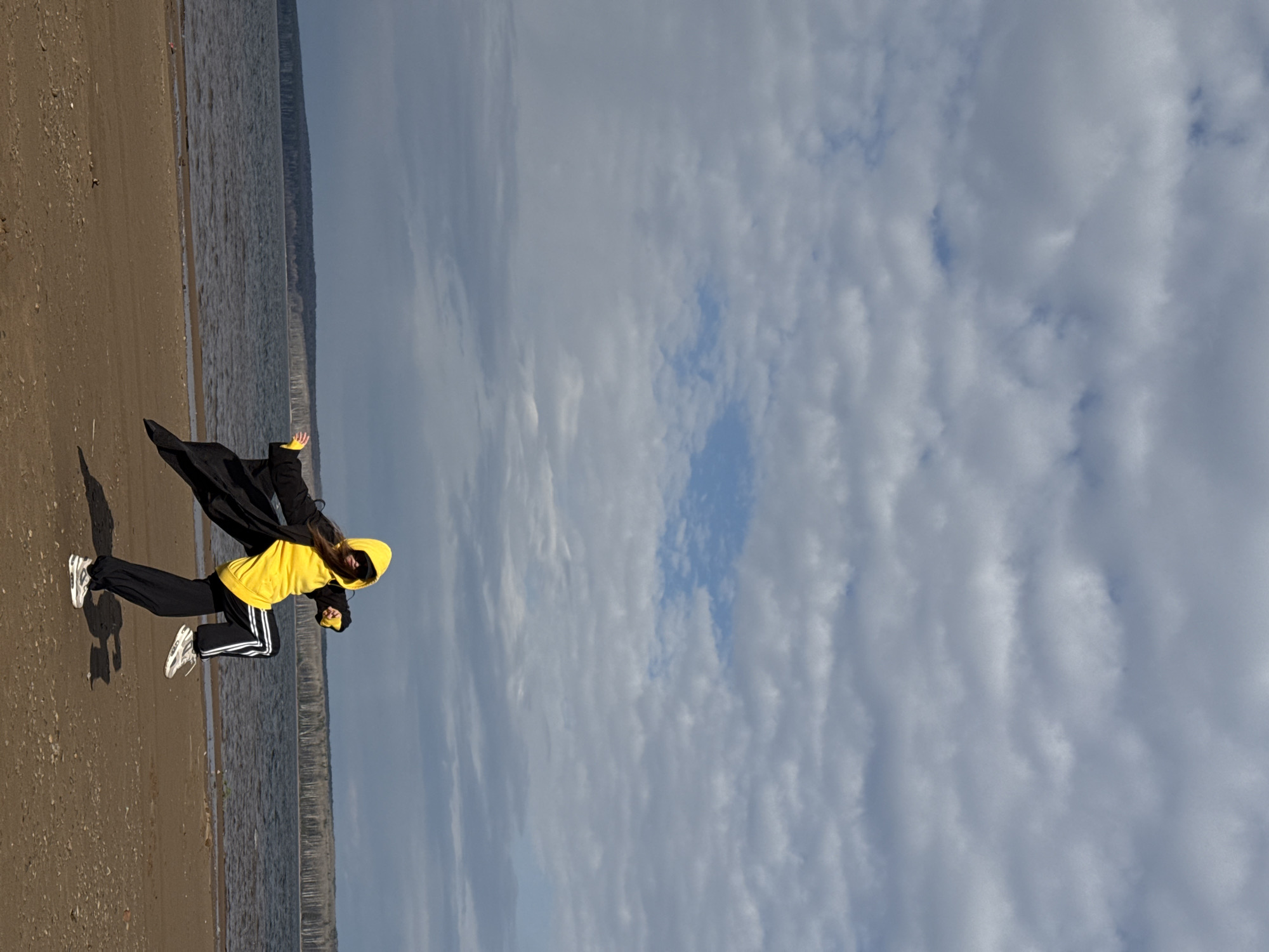 A person is captured in a dynamic pose on a sandy beach next to a wide body of water, likely a river or large lake, under a partly cloudy sky. The individual, appearing to be female, is wearing a black long coat, a bright yellow hooded sweatshirt, black track pants with three white stripes down the side, and white athletic shoes. She is in mid-stride or a dramatic pose, leaning forward with arms extended, her dark hair visible beneath the yellow hood. A distinct shadow of the person stretches across the sand to her left.
The setting appears to be a natural outdoor environment, possibly a riverbank or lakeside in Chaikovsky, Russia, given the context. The water extends into the distance, meeting a low, hazy horizon where a distant landmass or shore is faintly visible. The beach itself is a mix of sand and small pebbles. The sky is predominantly covered with layered grey and white clouds, though patches of clear blue sky are interspersed. The lighting suggests it is daytime, possibly late morning or early afternoon, with diffused sunlight contributing to the soft shadows. The dynamic pose suggests energy or playful movement. No discernible text is visible on any objects or clothing in the image.