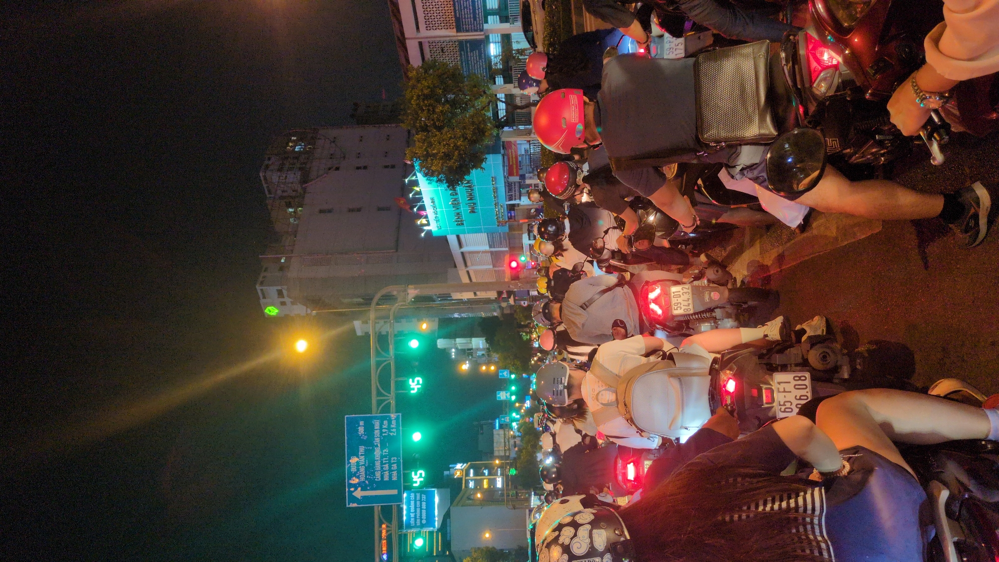 The image captures a bustling night scene on a city street, likely an intersection, filled with numerous motorbikes and their riders. The perspective is from a low angle, seemingly from a motorbike in the middle of the traffic, looking forward.
The primary subjects are many individuals on motorbikes, all wearing helmets of various colors and designs, indicating a common mode of transport. The motorbikes are densely packed, suggesting heavy traffic, and their brake lights illuminate the dark asphalt. Several traffic lights are visible, with green lights active for some lanes and red for others, indicating that vehicles are currently stopped, waiting for the light to change. A green digital display shows the number "45", likely a countdown timer for the traffic light.
The scene takes place at night, characterized by the dark sky, artificial street lighting, and the glowing red taillights and green traffic signals. In the background, multi-story buildings with illuminated windows are visible, along with trees.
Notable details include a large blue road sign on the left providing directions and distances. The sign reads "HOÀNG VĂN THỤ 500 m," "CẢNG HÀNG KHÔNG TÂN SƠN NHẤT" (Tan Son Nhat International Airport), "NHÀ GA T1, T2 - 1.9 Km," and "NHÀ GA T3 - 2.6 Km." This indicates proximity to the airport in Ho Chi Minh City. Another sign visible on a building reads "BỆNH VIỆN ĐA PHÚ NHUẬN" (Phú Nhuận General Hospital), directly identifying the area as Phú Nhuận District in Ho Chi Minh City, rather than Thủ Đức, as provided in the context. License plates such as "59-01 844.32" and "65-F1 96.08" are visible on some motorbikes. Another text snippet "LIÊN HỆ QUẢNG CÁO 0909 809 737" is also seen on a smaller sign. The overall impression is a typical busy night in a Vietnamese metropolis.