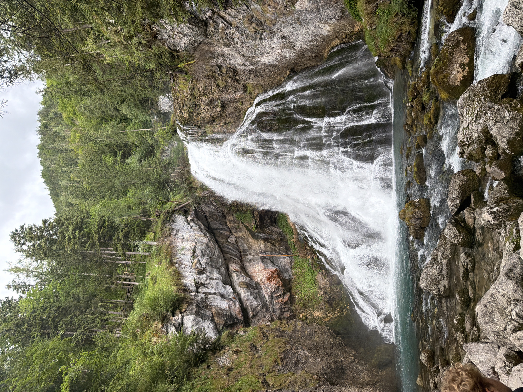 This image captures the Golling Waterfall in Golling an der Salzach, Austria. A powerful, multi-tiered waterfall cascades down a rocky cliff face covered in lush green vegetation. The water is a vibrant turquoise blue as it pools at the base, flowing over numerous rounded rocks and pebbles. Dense, green coniferous trees form a forest on the surrounding slopes, reaching towards a cloudy sky. In the foreground, a person with curly blonde hair is visible from the back, sitting amongst large grey rocks near the water. The scene is one of natural beauty and serene wilderness.
