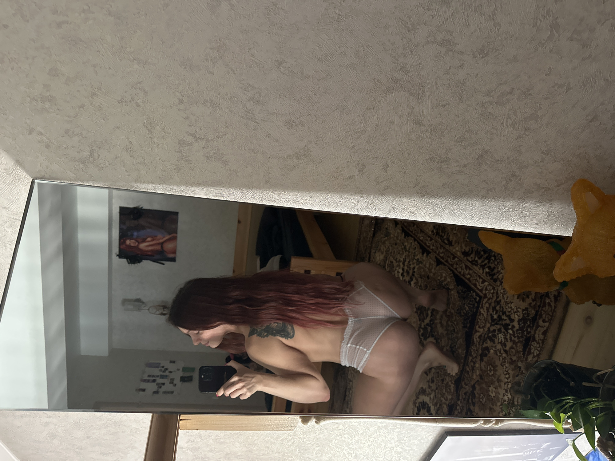 The image is a mirror selfie taken indoors, showing a woman with long, wavy reddish-brown hair. She is wearing light-colored, possibly white, sheer underwear with a dotted pattern, and appears to be bare-chested. A dark-colored tattoo is visible on her upper back or shoulder. She is holding a black smartphone in her left hand, suggesting she is taking the photograph herself. Her body is bent forward, and she is looking towards the phone screen.
The mirror reflects parts of an interior room. A patterned, dark-toned rug or carpet covers the floor. On the wall behind the woman's reflection, there is a framed picture depicting a woman with reddish hair in a reclined pose. Further to the right, a bright orange or yellow plush toy, possibly shaped like an animal, is partially visible. A green plant in a pot is also seen in the lower right corner of the reflection. The walls have a light-colored, textured wallpaper. The lighting suggests an indoor setting, possibly with artificial light. No explicit text is visible in the image. The scene conveys a personal and intimate moment within what appears to be a home in Vladimir, Russia.