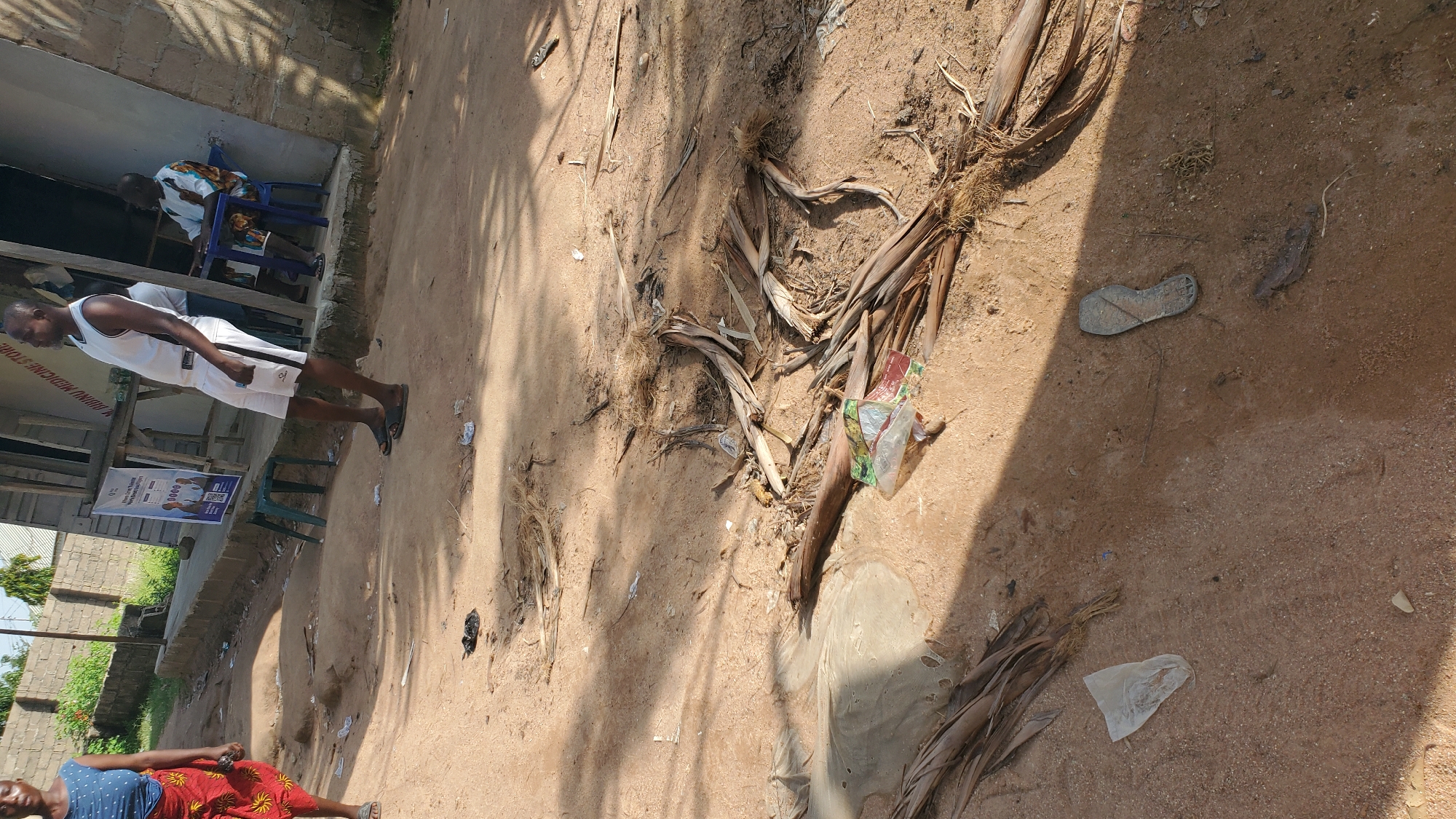 The image depicts a street scene in Jalingo, Nigeria, characterized by a dirt road and a sandy ground surface. In the foreground, palm fronds and various debris are scattered across the ground, including a plastic bag and a single shoe sole. Further back, a woman wearing a blue sleeveless top and a red patterned skirt stands on the left, facing away from the camera. To the right, a man in a white tank top and white shorts stands facing left, with his back partially to the camera. He is positioned near the entrance of a building. Another individual, a man in traditional attire with a colorful patterned top, is seated on a blue chair to the right, inside the building's entrance. A table is positioned in front of him. The building appears to be a shop, indicated by signage that includes "M. JABINU MEDICINE STORE." A poster displaying product information and possibly advertisements is attached to the front of the building. Two green plastic chairs are placed on the ground in front of the building. The overall environment suggests an outdoor commercial area in a town or city.