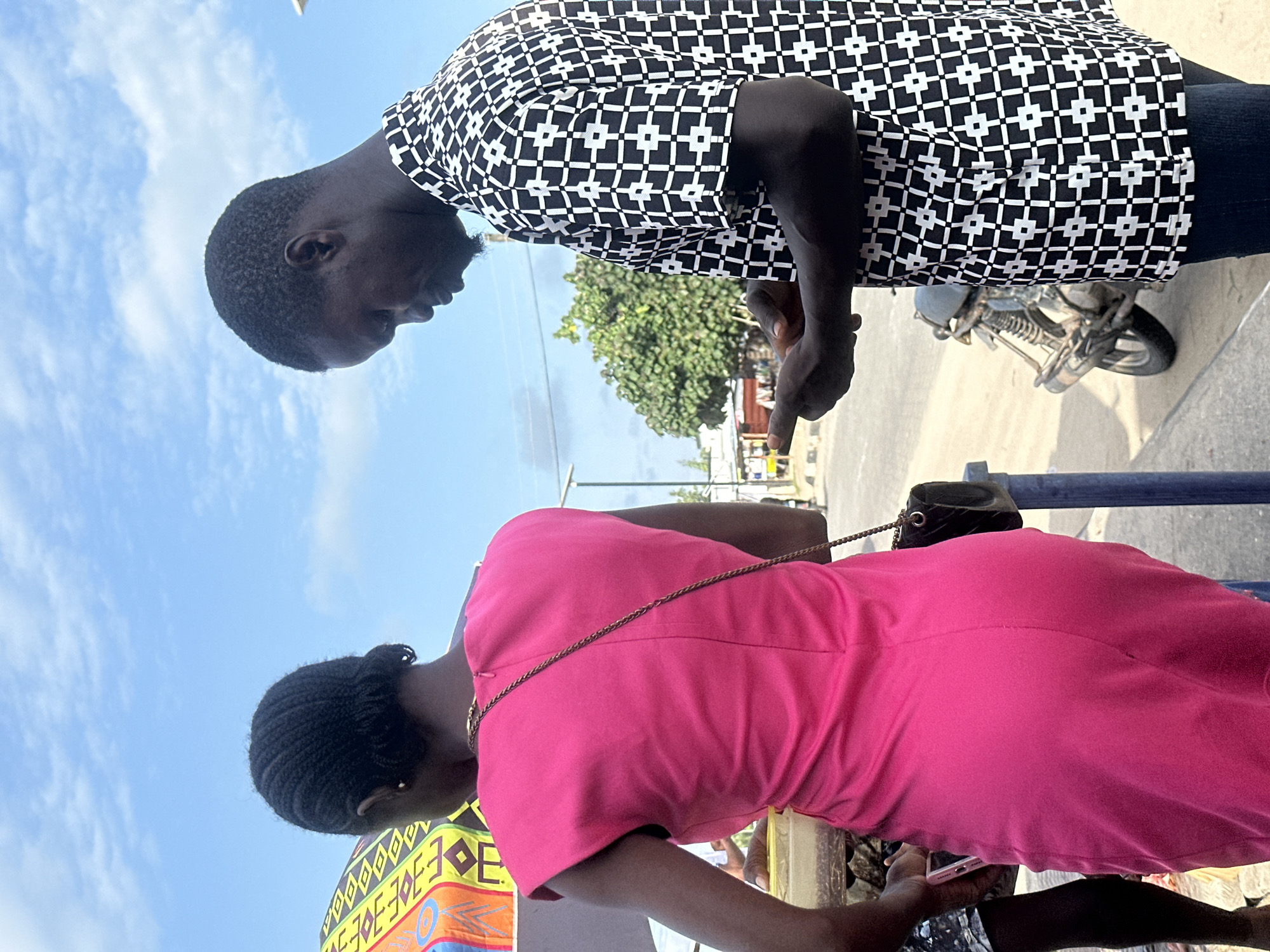 Two people, a man and a woman, are standing outdoors. The man, wearing a black and white patterned shirt, is looking down and to his right, possibly at a phone held by the woman. The woman is wearing a bright pink dress and is also looking down, seemingly engaged with the phone. A small black purse with a chain strap is visible on her shoulder. In the background, a motorcycle is parked. The scene appears to be a bright, sunny day with a clear blue sky and scattered clouds. The presence of the motorcycle and the general outdoor setting suggest a casual, everyday environment. The overall mood seems relaxed and focused on the interaction between the two individuals. There is no visible text in the image.