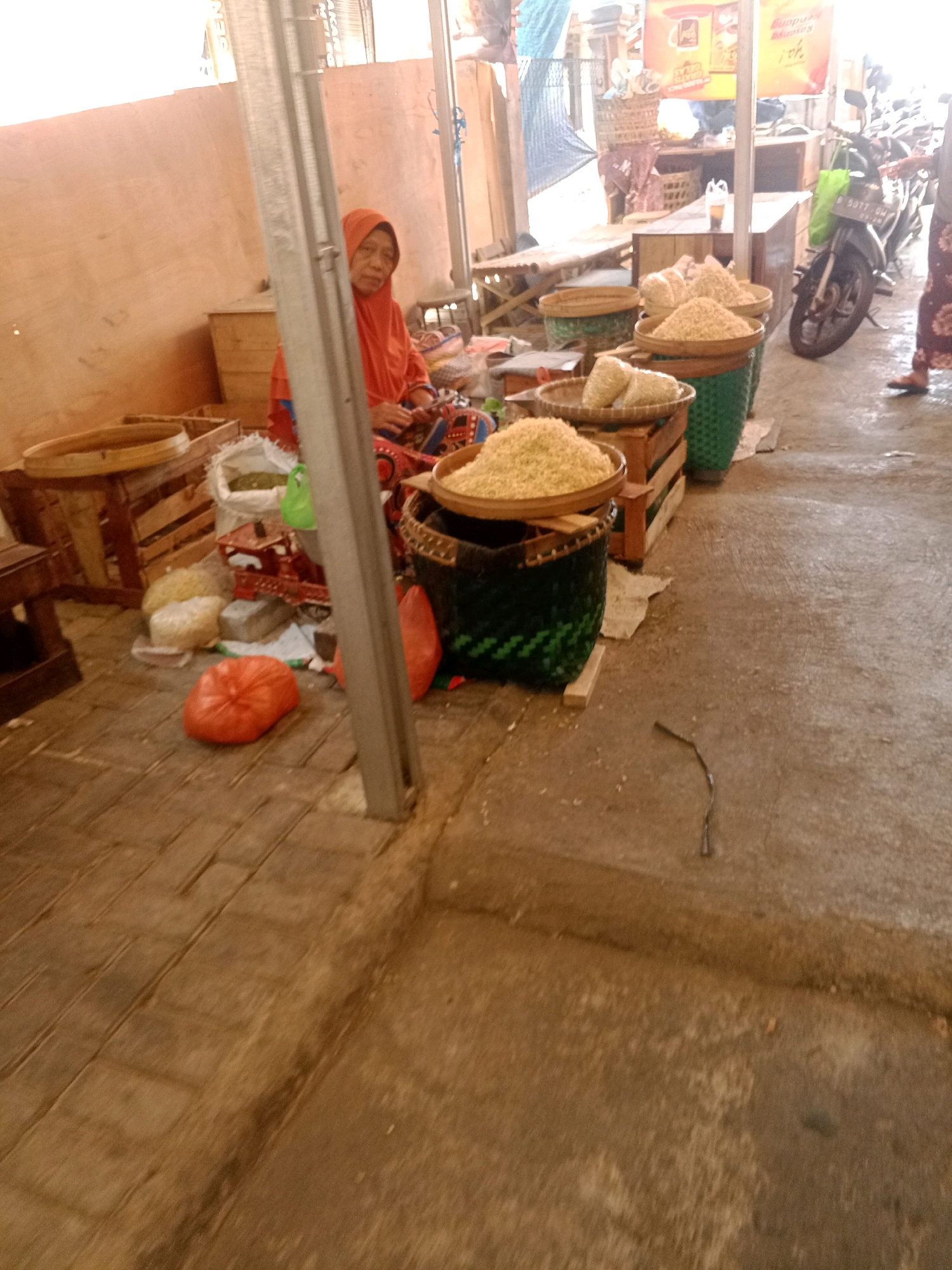 A woman wearing an orange headscarf and a patterned dress sits behind a stall at an outdoor market in Wonogiri, Indonesia. The stall is filled with large baskets of grains, possibly rice or corn, and other produce. In the foreground, an orange bag sits on the ground. A motorcycle is parked to the right of the stall, and a person's legs are visible near it. The ground is paved with bricks and dirt. The stall is covered with an orange tarp. The image appears to be taken during the daytime, with bright sunlight casting shadows.