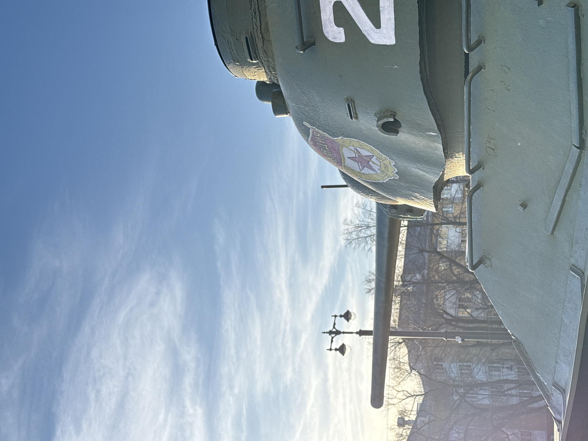 The scene is set outdoors in Perm, Russia. The primary subject is an olive-green military tank, viewed from its front-right quarter at a low angle. The tank's turret features a white numeral "2" and a distinct Soviet-era emblem comprising a red star within a wreath, surmounted by the Cyrillic word "ГВАРДИЯ" (Guard). The tank's long main gun barrel extends horizontally towards the left of the frame. The tank's surface appears textured and slightly worn. In the background, behind the tank, an ornate dark streetlamp with two illuminated globes stands amidst a collection of bare deciduous trees. Further beyond the trees, multi-story residential buildings are visible. The upper portion of the image is dominated by a clear blue sky with thin, wispy white clouds. Soft, warm light suggests late afternoon or early morning. No people or dynamic actions are present in the image.