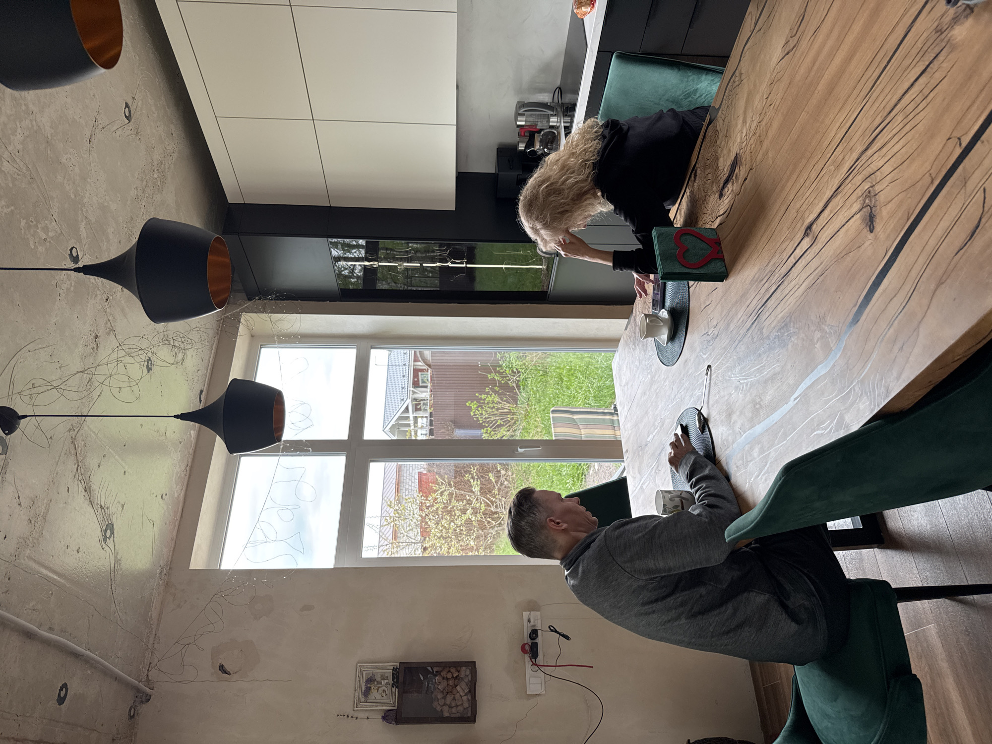The image depicts the interior of a room, likely a kitchen or dining area, in Steklyannyi, Russia. Two individuals are seated at a large wooden table. A woman with blonde hair is seated on one side, facing toward the window, and she appears to be looking at or interacting with something on the table. A man in a grey sweatshirt is seated opposite her, also looking down at the table. The table is set with placemats and cups.
The room features a large window providing a view of a residential area outside. Visible outside are houses, trees, and greenery.
The interior design includes black pendant lights with gold interiors. The walls are partially decorated with exposed wiring and artistic embellishments. The room also includes a kitchen area with modern cabinets. On the wall is what appears to be framed artwork.