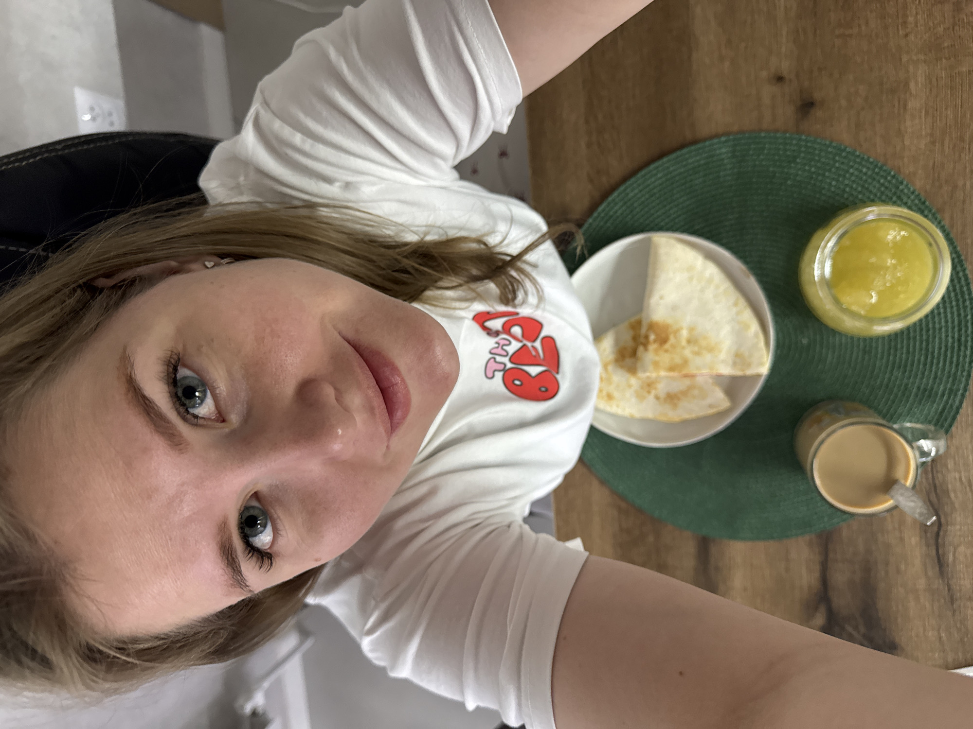 The image shows a selfie of a woman with light skin and blue eyes, wearing a white t-shirt with a cartoon design. She is positioned above a table setting. On the table, there is a plate containing quesadillas, a jar of what appears to be yellow sauce or spread, and a mug of coffee with a spoon. These items are placed on a green placemat. The table is made of wood. The location is indoors, likely a dining area. There is no evidence to confirm the location of Gornyi (Dvurechenskoe S/P), Russia.