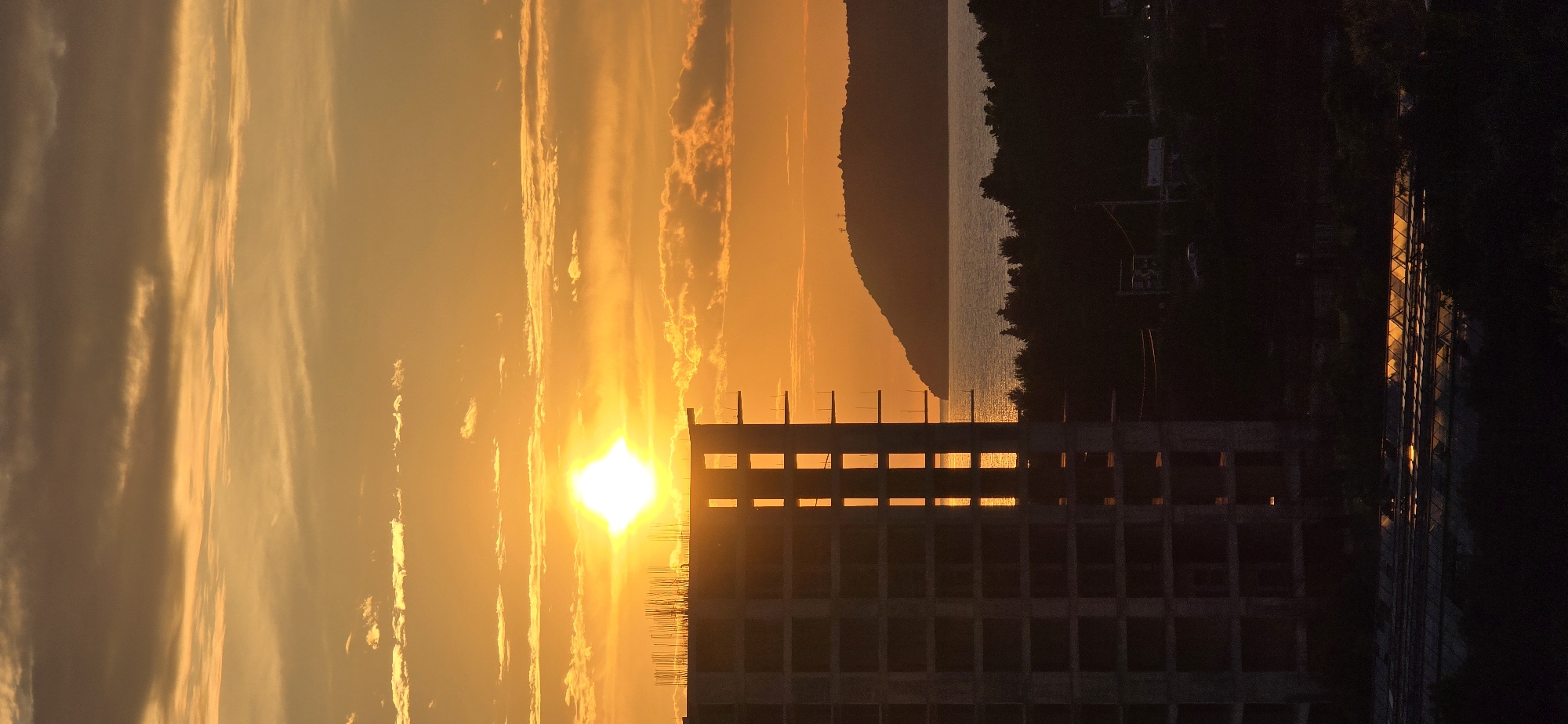 A vertical landscape photo shows a sunset obscured by a building in Bar, Montenegro. The sun is bright white-yellow, shining through light, vertical, orange clouds. The unfinished, rectangular building in the foreground has open windows. The background shows a sliver of water and a dark, forested mountain range. To the right of the building are trees and a building with orange lights.
