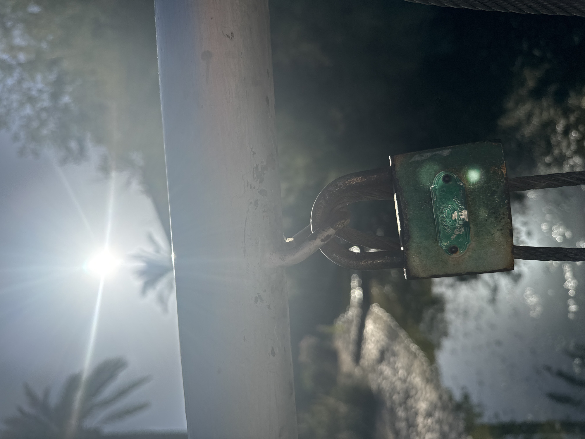 The image captures a close-up of a green, rusted padlock hanging from a metal railing. The padlock is old and shows signs of wear and tear. Behind the padlock, the sun is prominently visible, creating a bright flare and casting lens flares across the image. The background is out of focus, showing a blurry representation of water with shimmering reflections and some foliage. The overall lighting suggests it is daytime, possibly morning or late afternoon, with the sun positioned high in the sky. The context of Murcia, Spain, is not visually evident from the image itself.