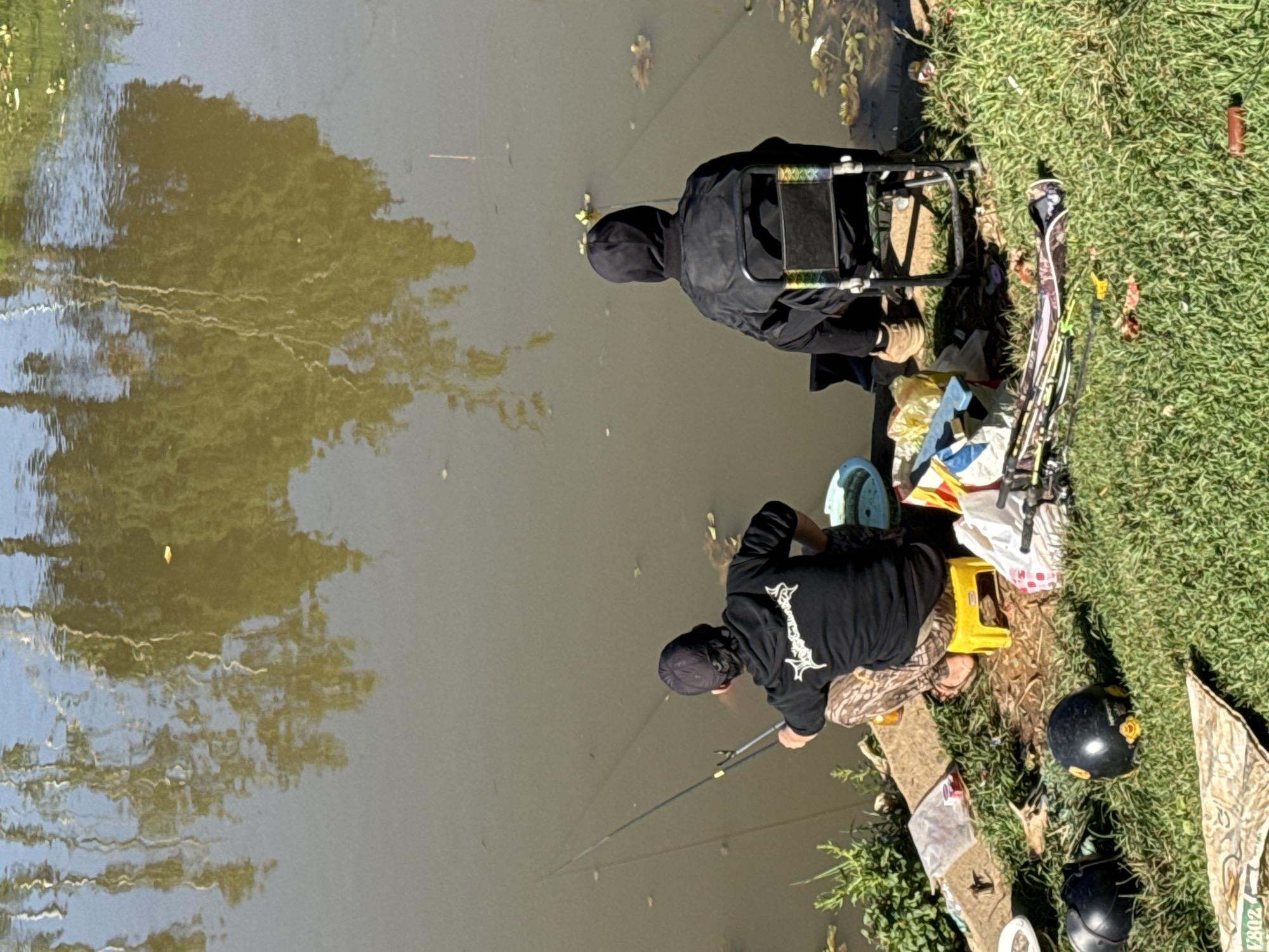 The image captures an outdoor scene of two individuals fishing by the edge of a murky body of water, likely a pond or river, in Da Lat, Vietnam. The time of day appears to be sunny, with clear skies reflected in the water, indicating good weather.
In the foreground on the right, one person is seen from behind, hunched over a fishing rod extending into the water. This individual wears a black long-sleeved shirt with a distinctive white graphic print on the back, possibly stylized letters, and camouflage-patterned pants. They are seated on a small yellow plastic stool. Next to them on the grassy bank are various items including bags, possibly containing fishing bait or equipment, and two dark-colored helmets, which resemble motorcycle helmets. A camouflage patterned cloth or bag nearby shows partial text, "202".
Further down the bank, slightly behind the first person, another individual is also engaged in fishing. This person is dressed entirely in black, including a hooded jacket that obscures their face, and sits on a black folding chair. Their fishing rod is also cast into the water.
The bank is covered in green grass, with some scattered leaves and small pieces of debris. The water itself is a dull brownish-green, reflecting the bright sky and the dense foliage of trees along the opposite bank or overhead. These reflections create wavy patterns on the water's surface. The overall atmosphere suggests a calm and focused activity amidst a natural setting.
