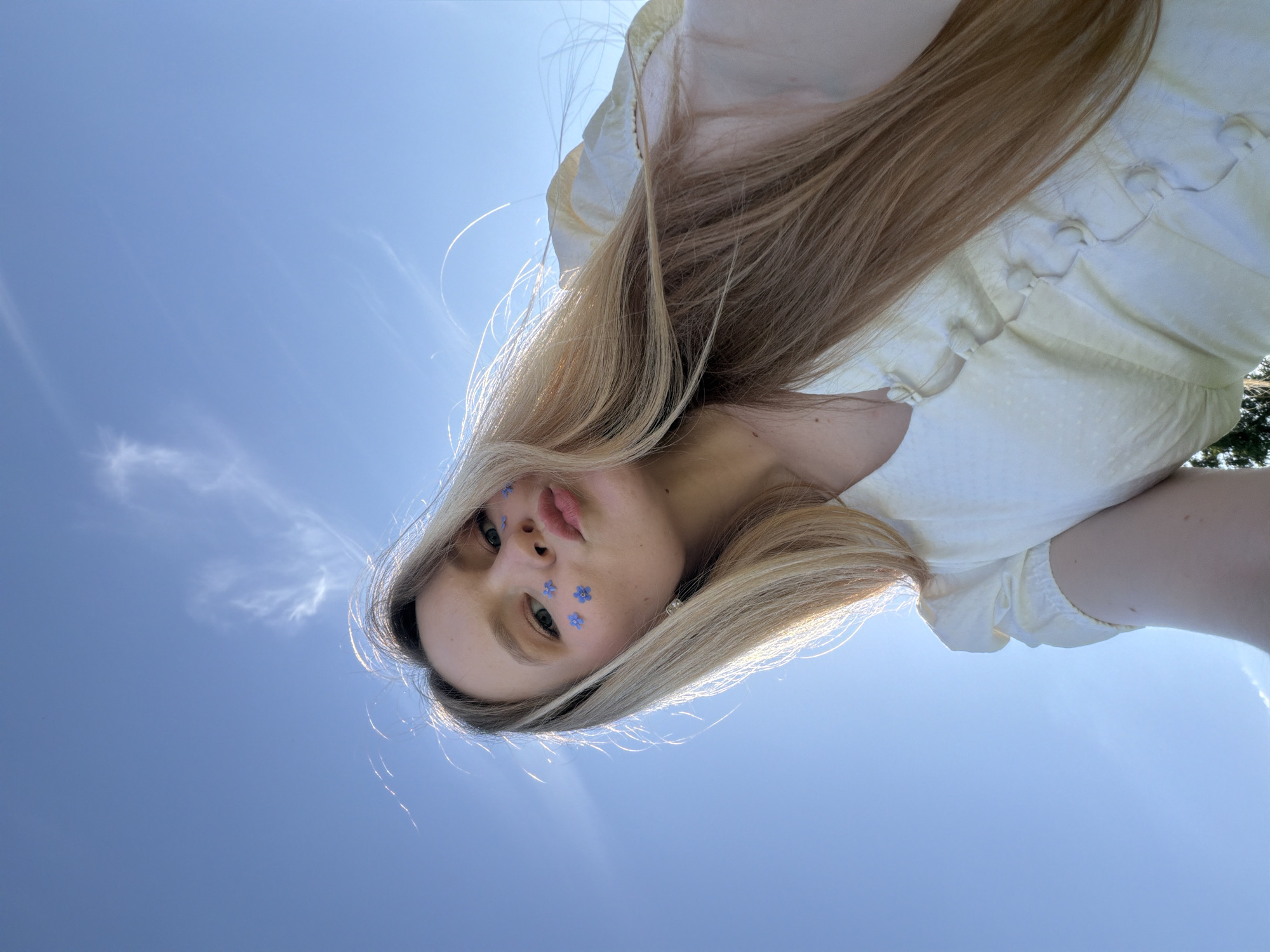 A woman with long, blonde hair is looking down at the camera from a low angle. She is wearing a light-colored top with small buttons down the front. There are several small, blue flower stickers on her face. The background is a clear blue sky with some wispy clouds. The location, Кадряково, Russia, cannot be determined from the visual information in the image.