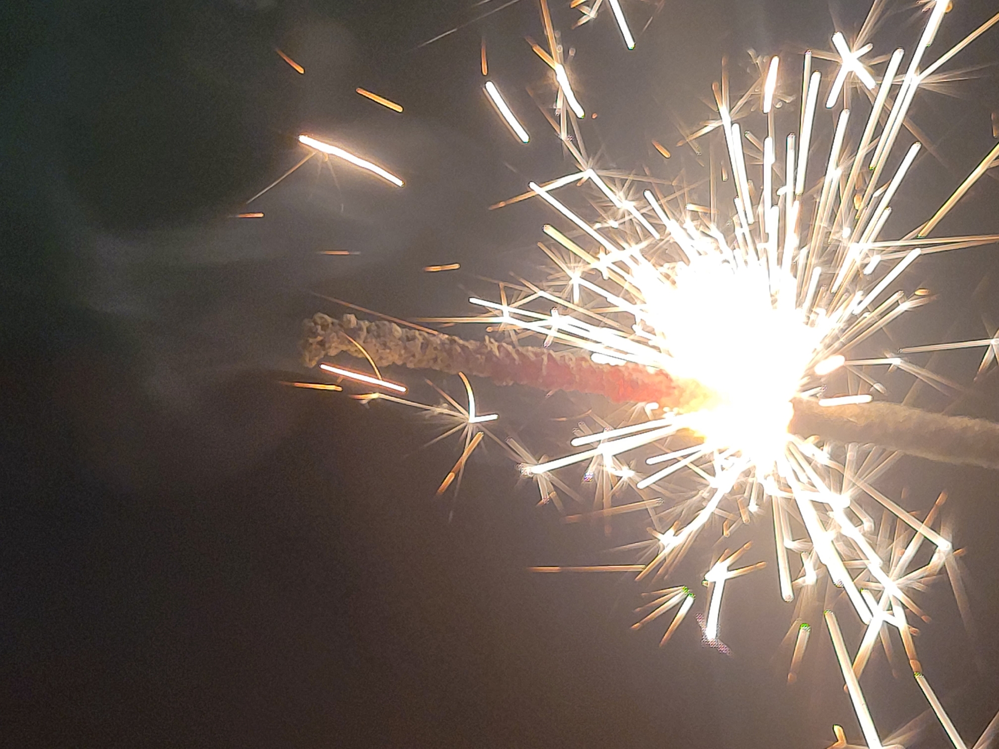 The image shows a sparkler burning against a dark background. The sparkler is mostly consumed, with bright, white-yellow sparks radiating outward from the point of combustion. Some embers are visible, and a trail of smoke extends from the burning end of the sparkler. The background is dark, with no other objects or details visible. There is no information in the image to determine any location, much less the city of Izhevsk, Russia.
