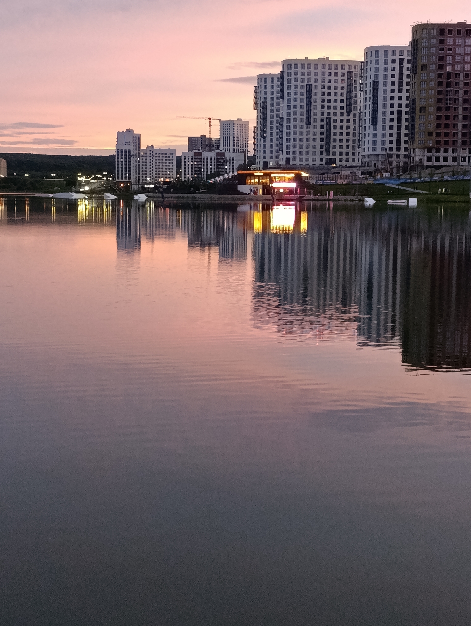 The image depicts a waterfront scene at dusk in Penza, Russia, specifically along the embankment of the Sura River near the city center. In the foreground, the calm water of the river reflects the sky and the buildings on the opposite bank. The sky is painted with soft hues of pink and purple, characteristic of a sunset.
On the far bank, a row of modern, high-rise residential buildings stretches across the horizon. Some of these buildings appear to be completed, with their windows reflecting the fading light, while others are still under construction, showing exposed concrete and structural elements. A prominent feature is a building with a lit facade, possibly a restaurant or cafe, situated at the water's edge, its golden glow vividly mirrored in the river. Behind the buildings, a line of trees and streetlights can be seen, indicating the proximity of the city's infrastructure. The overall impression is one of urban development meeting natural landscape, with the tranquil water serving as a reflective surface for the city's evolving skyline.