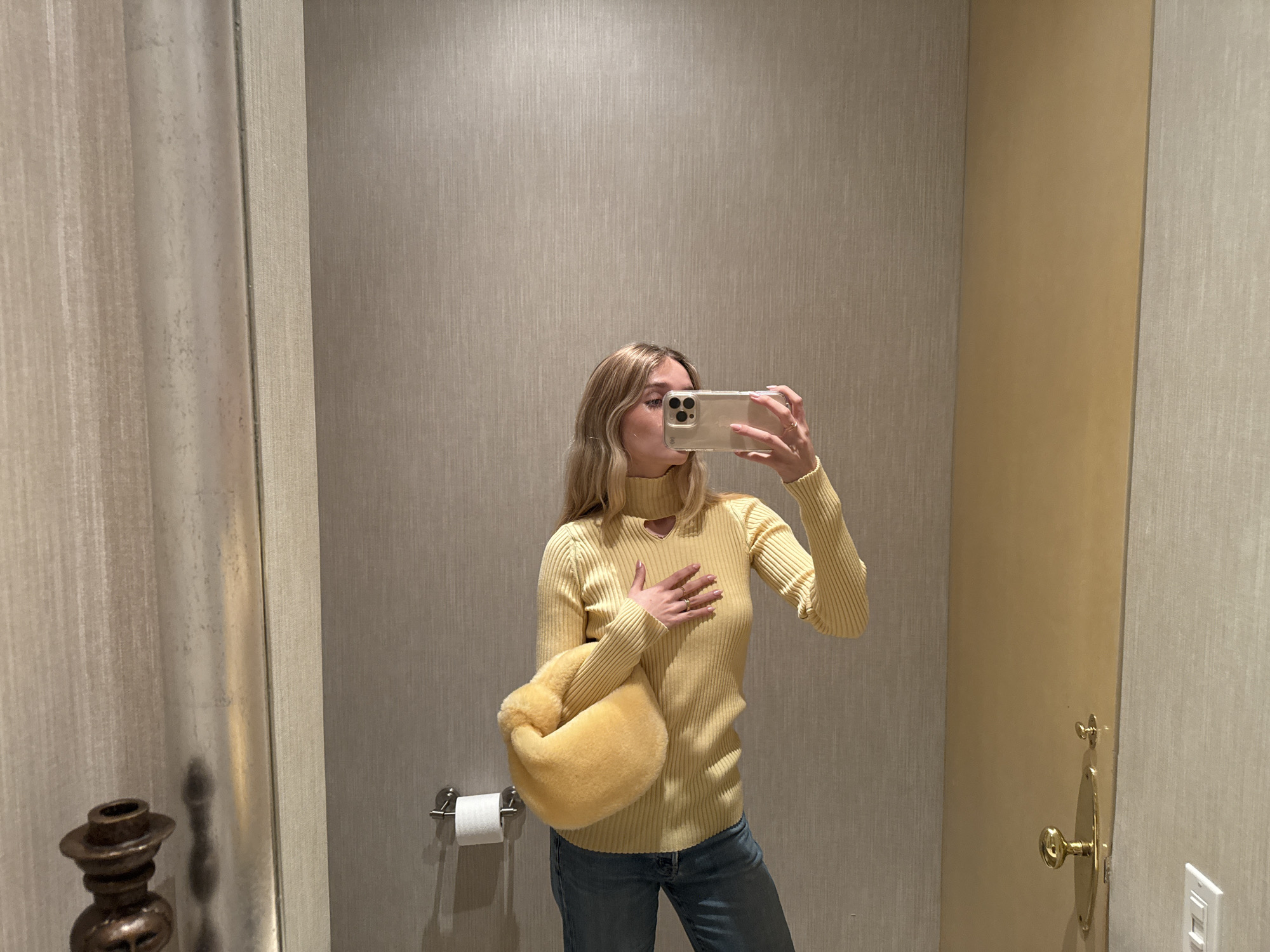A fair-skinned woman with blonde wavy hair is taking a selfie in a restroom in Mexico City, Mexico. She is wearing a light yellow ribbed turtleneck sweater with a cutout at the neckline, blue jeans, and is carrying a fluffy yellow purse in her left arm. She is holding a light-colored iPhone in her right hand, angled to capture her reflection in a mirror. The restroom has walls with vertical textured wallpaper in beige and gray. A metal toilet paper holder with white toilet paper is visible next to the woman's left side. A decorative metal object is visible in the bottom left of the image. A door with gold-colored hardware and a white electrical outlet is visible to the woman's right.