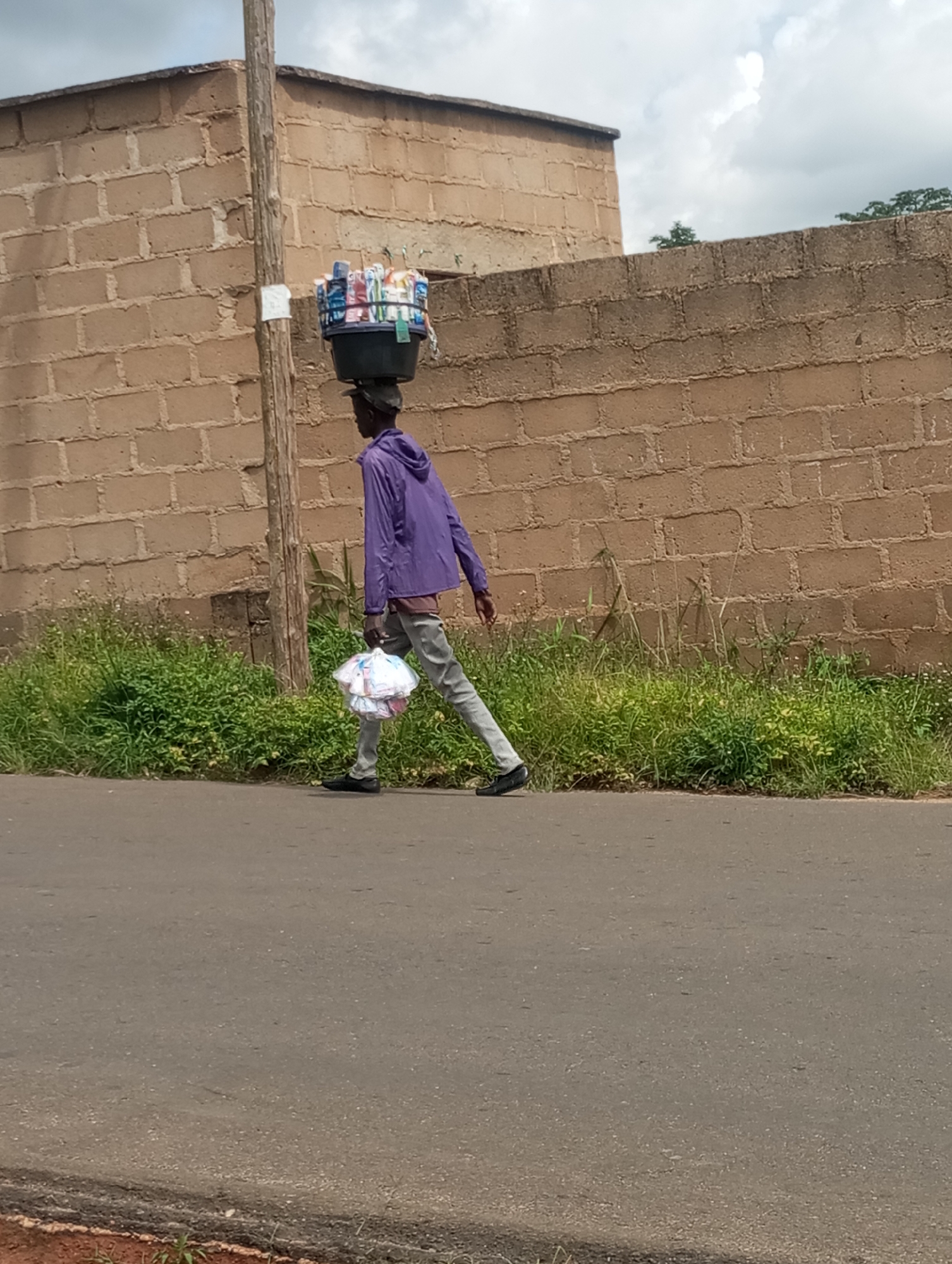 The image captures a side view of a young Black person walking on a paved road. The individual is carrying a black basin filled with assorted items on their head and a white plastic bag in their right hand. They are wearing a purple hooded jacket and light gray trousers. The background features a brick wall and dense green vegetation. The scene appears to be located in Jos, Nigeria, a city known for its brick-making industry and its position on the Jos Plateau. The sky is overcast with clouds.