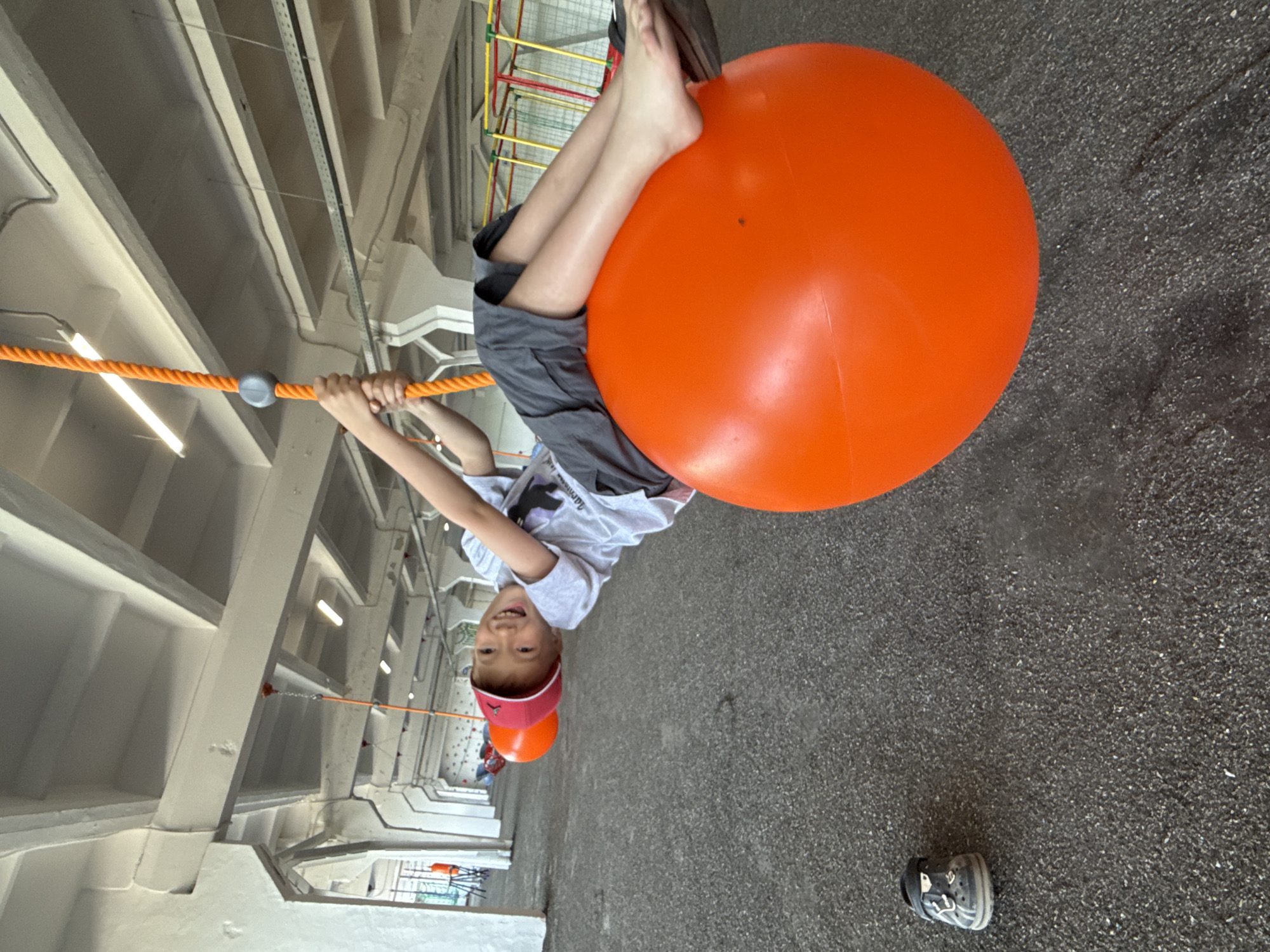 A child is hanging upside down from a rope swing, using an orange exercise ball as a seat, at an indoor playground. The child is gripping the rope with their hands, their legs are extended over the ball, and they are smiling at the camera. The playground appears to be located inside a building with a high ceiling with visible pipes and structural elements. A single shoe is lying on the ground near the edge of the frame. The photo was taken in Sin'kovo, Russia.
