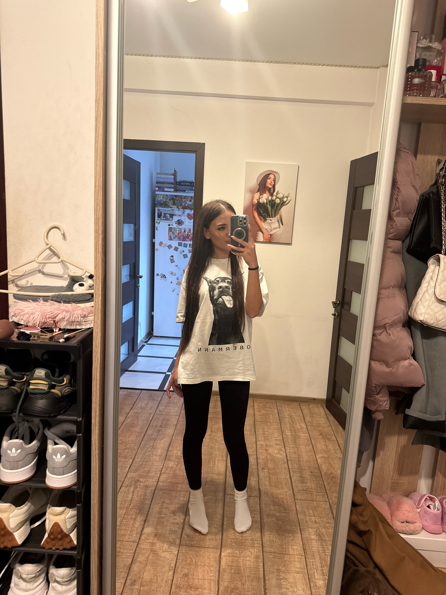 The image is a full-body selfie taken in a room, likely a bedroom or entryway, in Chișinău, Moldova. A young woman with long dark hair is standing in front of a large mirror, holding a smartphone to take the picture. She is wearing a white t-shirt with a black graphic of a dog and black leggings. She also has white socks and white pointed-toe ankle boots.
The room appears to be a home setting. To the left of the woman is a tall, narrow shoe rack filled with various shoes and a clothing rack with a fluffy gray stuffed animal and a pink accessory hanging from it. Behind her, a door is visible, and further in the background, a refrigerator is adorned with magnets and photos, suggesting a kitchen area. The floor is covered with wooden laminate flooring.
To the right of the woman, reflected in the mirror, is a tall, floor-to-ceiling mirror that occupies a significant portion of the frame. Behind the mirror, a closet or wardrobe area is visible, with coats and handbags hanging on hooks. On a shelf above the coats, several bottles of perfume or toiletries are displayed, along with a framed picture of a woman holding tulips. The overall impression is of a lived-in personal space.