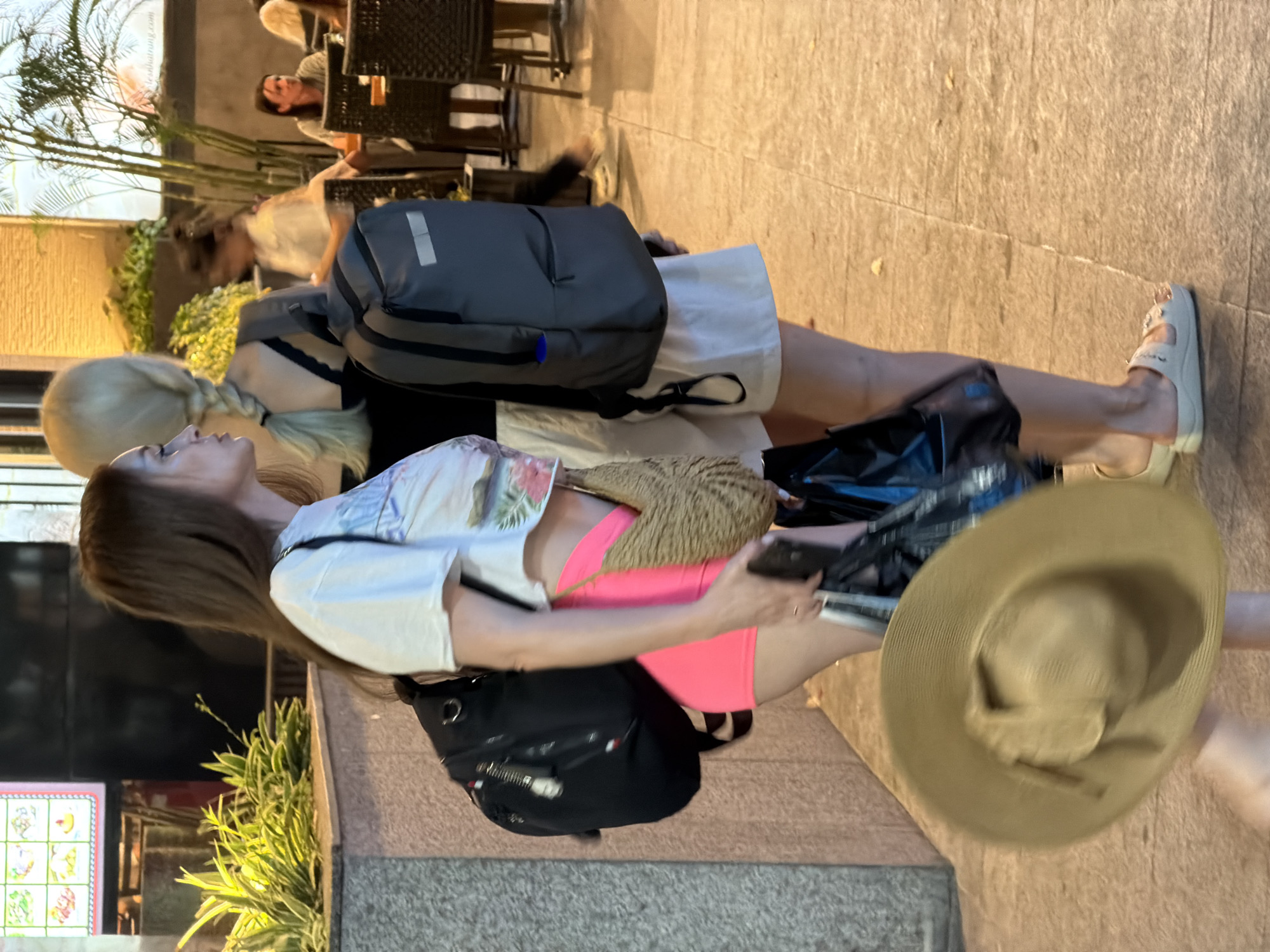 The image shows a vertical full shot of several people and objects on a paved walkway outside at what appears to be dusk or early evening, given the artificial lighting. In the foreground, a woman with long brown hair is seen from the side, wearing a white t-shirt with a floral print, bright pink shorts, and carrying a black backpack. She is holding a phone in her right hand. To her left and behind her, another person with blonde hair in a braid is partially visible, also with a backpack. A large, straw-like sun hat is also prominent in the foreground, obscuring some of the scene.
Behind the foreground subjects, a man with a grey backpack is walking. In the background, there are outdoor seating areas with chairs and tables, suggesting a cafe or restaurant. Lush green plants are visible along the walkway. The overall setting appears to be a well-lit urban area in Nha Trang Ward, Vietnam, possibly a tourist or commercial district. The activity appears to be people walking or moving through the area, possibly after a day out. There are no explicit emotional cues, but the lighting and the casual attire suggest a relaxed, possibly warm evening. There is no visible text in the image.