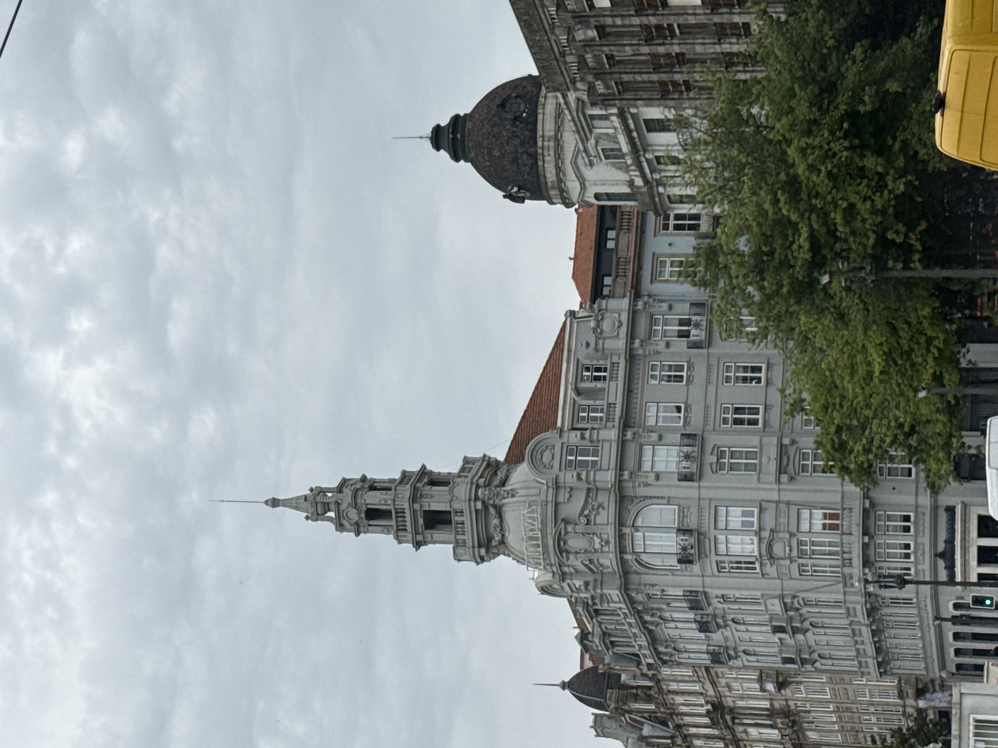 This is a vertically oriented medium shot of a European street scene with ornate buildings under an overcast sky. The architecture features intricate detailing, including columns, balconies, and mansard roofs, with a prominent domed structure and a tall, spire-like turret on one of the buildings. Lush green trees are interspersed between the buildings, adding a touch of nature to the urban environment. A portion of a bright yellow vehicle, possibly a van, is visible on the right side of the frame, adding a pop of color. The scene appears to be captured during the daytime, given the general illumination, but the cloudy sky suggests a muted, diffused light. There are no people visible in the frame, and no text is discernable. The style of the architecture hints at a historical European city, and the provided location context confirms this as Porto, Portugal. The overall atmosphere is one of a quiet, overcast day in a city rich with architectural heritage.