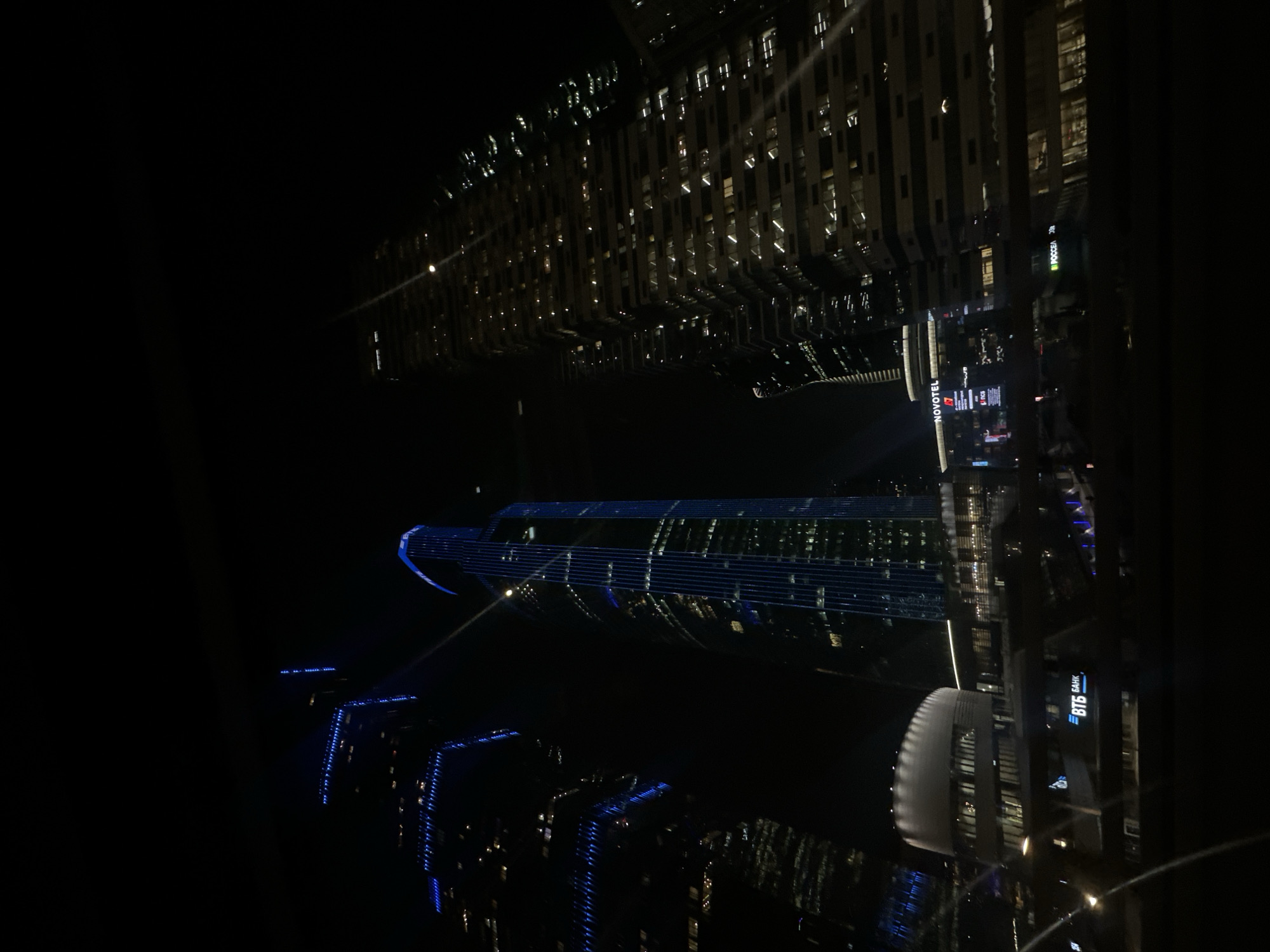 The image shows a nighttime view of buildings in Moscow, Russia, likely taken from a high vantage point or reflection. Notable buildings include the Novotel and VTB Bank, illuminated with lights. One building is lit in a blue hue. The environment is dark and urban, with a focus on the city's architectural elements at night.