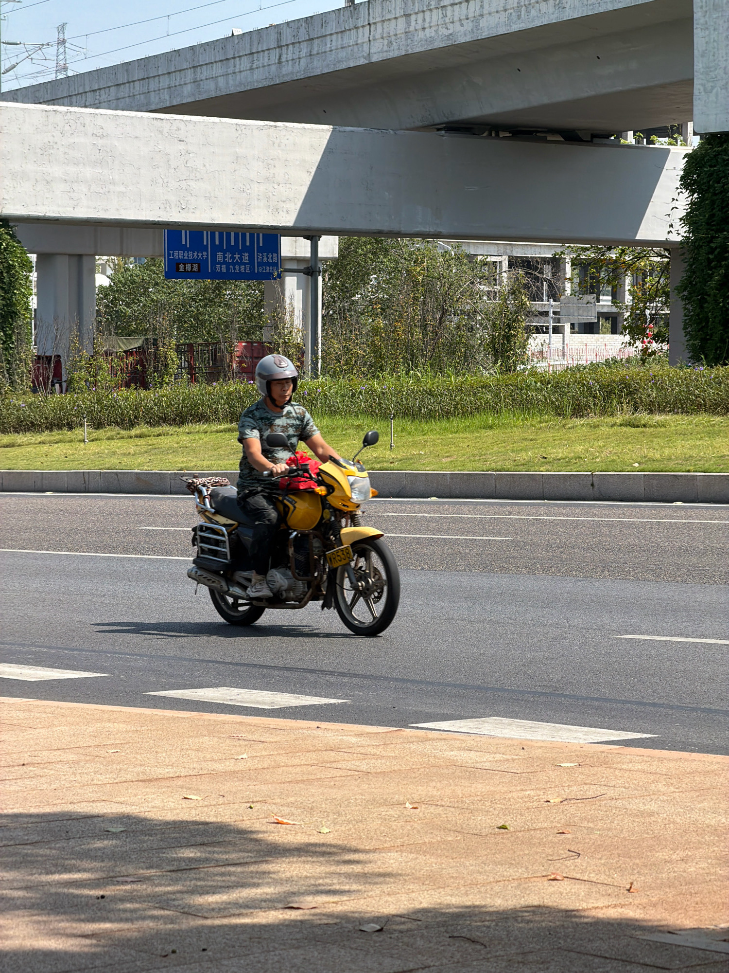 A man is riding a yellow motorcycle down a paved road in Jiangjin, China. He is wearing a camouflage t-shirt and a light gray helmet. The motorcycle has a license plate that reads "R538". In the background, there is a grassy embankment with some trees and shrubs, and a large concrete overpass. A blue directional sign is visible on the overpass, indicating directions to "South Avenue" and other locations. The sky is clear and blue. The road has white lane markings.