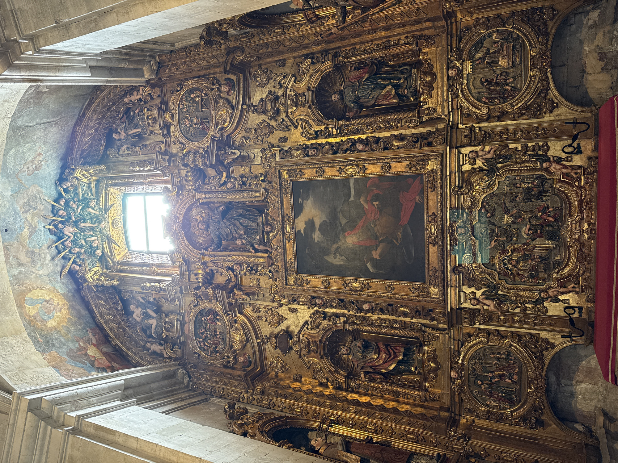 The image depicts the ornate ceiling of a church in Unknown, Unknown. The ceiling is heavily decorated with gold ornamentation, including sculpted figures, reliefs, and frames. A large, rectangular painting is centrally located. Niches contain statues of religious figures. The style is likely Baroque. A window is visible on the left.