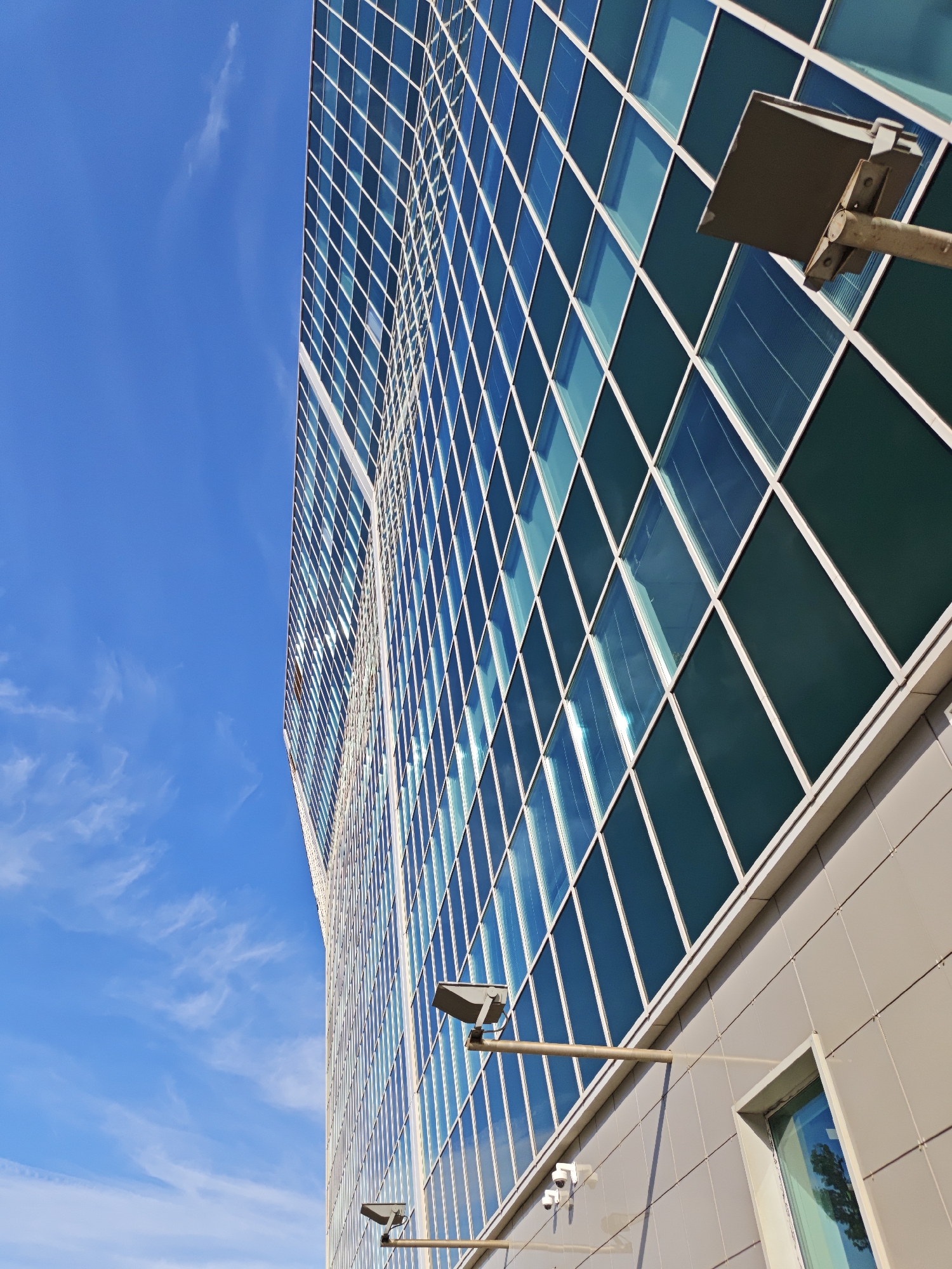 The image shows the exterior of a modern, glass-facaded skyscraper against a bright blue sky with wispy clouds. The building's facade is composed of numerous diamond-shaped panes of glass, some reflecting the sky and clouds, others appearing a deep, dark blue or green. The perspective is low-angle, looking upwards at the towering structure.
Two security cameras are mounted on horizontal arms extending from the building's exterior. One is larger and positioned higher up, while a smaller one is visible below it. A rectangular light fixture, also mounted on an arm, is attached to the building near the top of the frame. The lower portion of the building visible is clad in beige or light grey panels, with a framed window reflecting trees.
The scene suggests daytime, likely morning or afternoon given the bright sky and sunlight. The weather appears clear and sunny. While the specific location is not visually identifiable beyond being a modern urban environment, the context provided indicates Moscow, Russia. There is no text visible in the image. The overall impression is one of contemporary architecture and urban infrastructure.