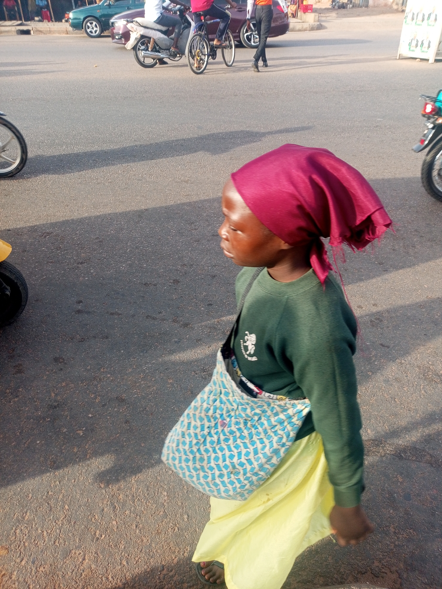 A young individual wearing a maroon headscarf, a long-sleeved green shirt with a logo, a light yellow skirt, and sandals, walks on a paved street. They carry a blue and white patterned shoulder bag. In the background, there are cars, motorcycles, and pedestrians. The location appears to be a city street.